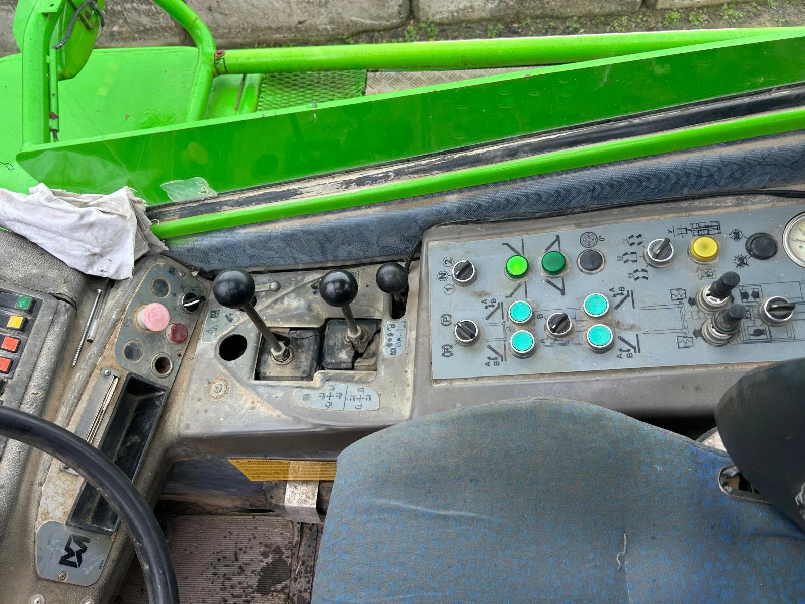 Close-up of the control panel, levers, and steering wheel inside a heavy machinery cab, with a green exterior frame visible through the window.