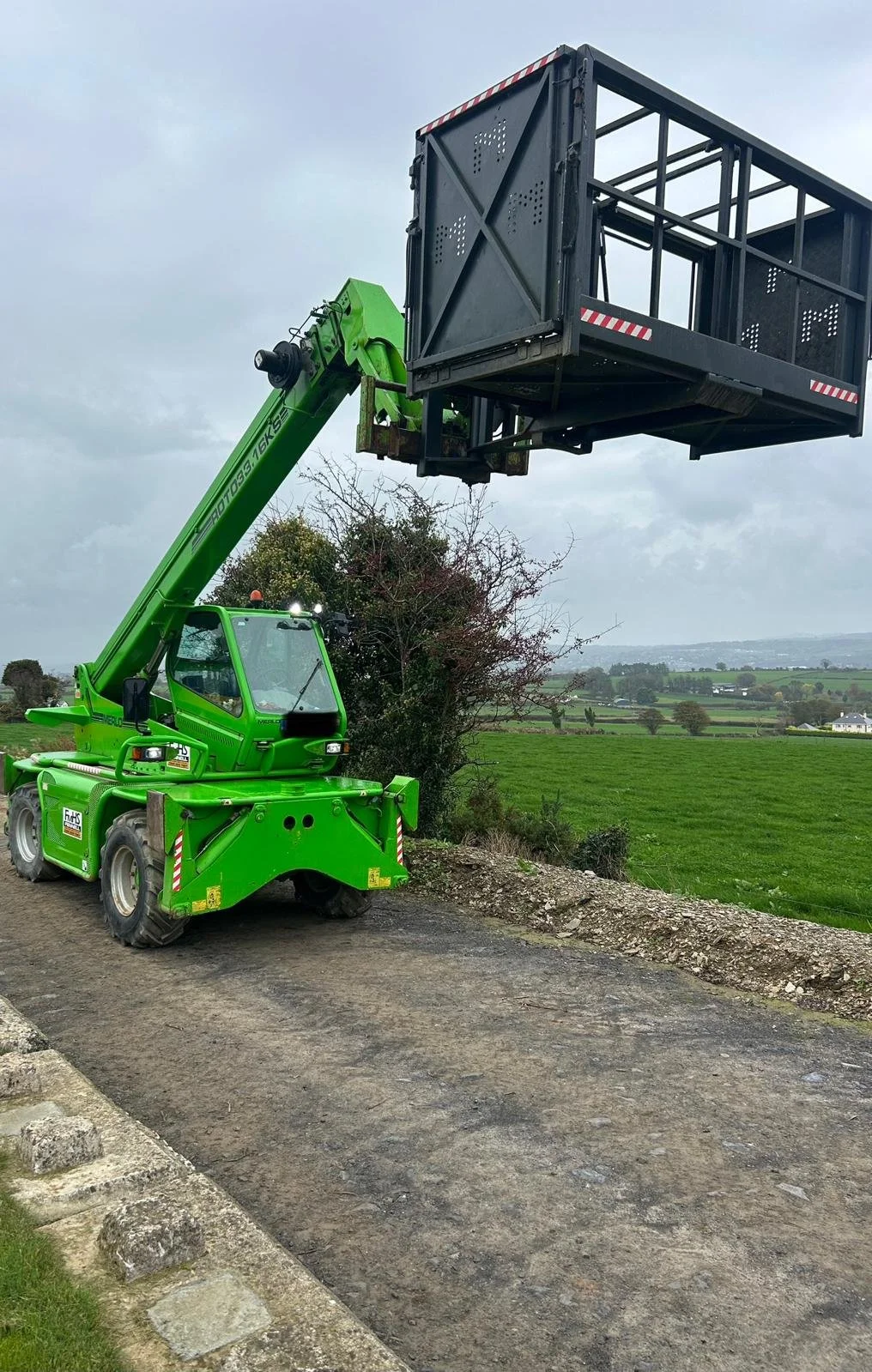 A green telescopic boom lift with a black work platform is extended high in the air, set against a cloudy sky. The lift is on a dirt path next to a grassy field with trees and rolling hills in the background.
