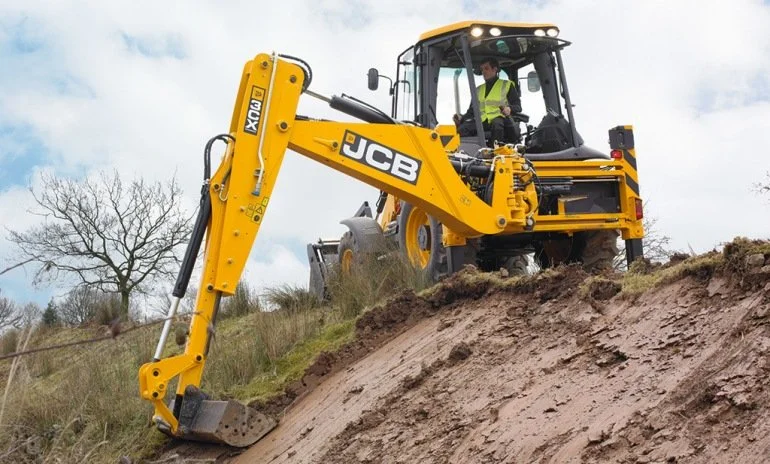 A yellow JCB excavator with a worker inside operating it, working on a slope outdoors under a cloudy sky.