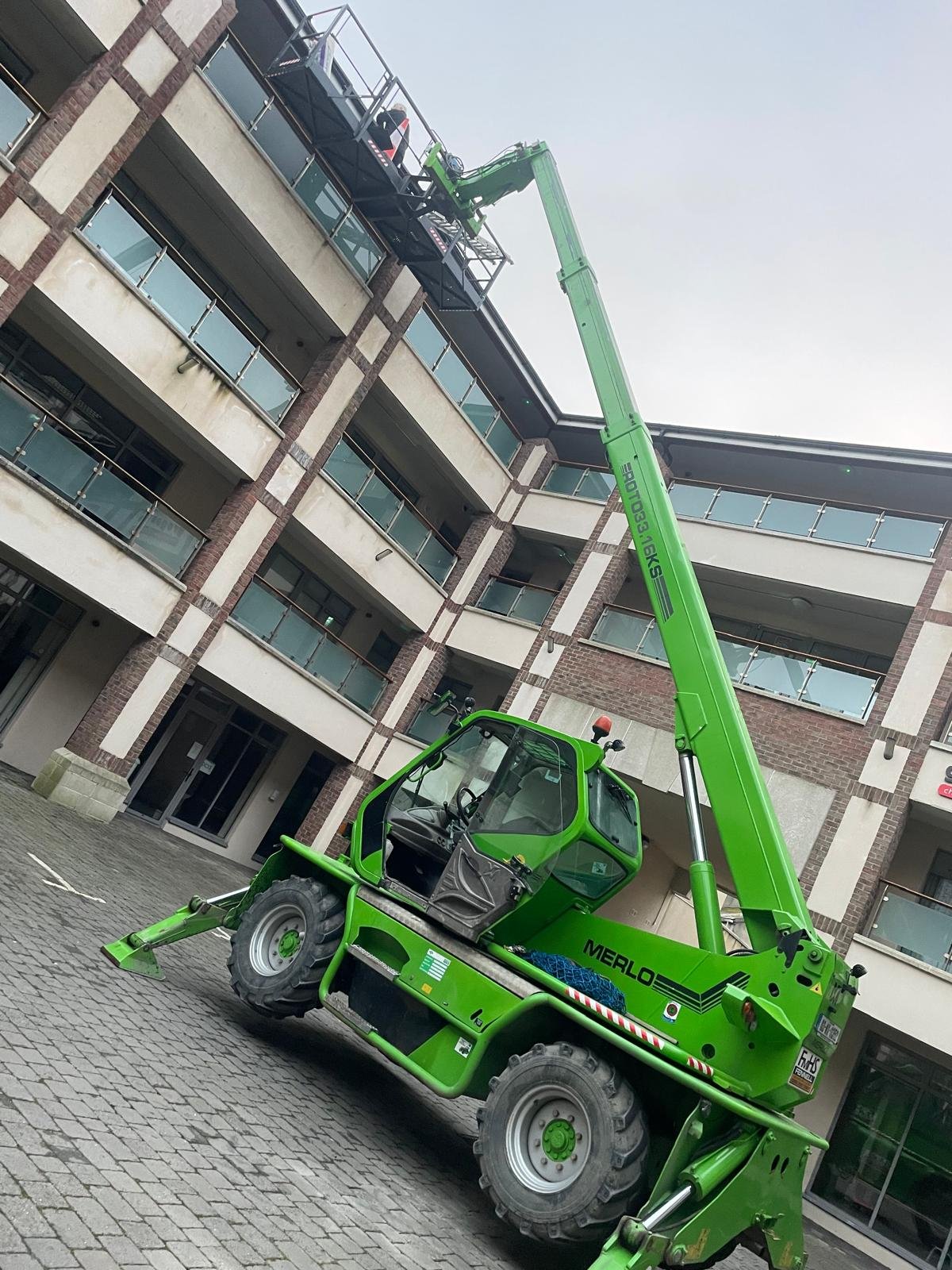 A bright green telescopic boom lift extending toward the upper floors of a multi-story building with balconies and glass railings.