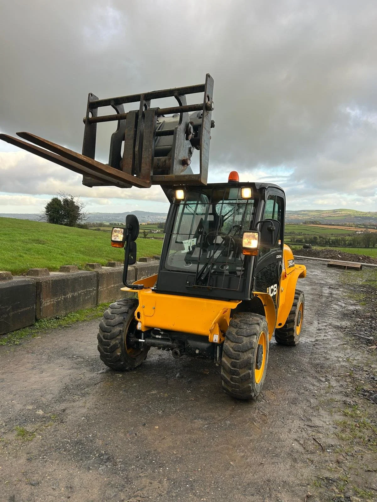 A yellow JCB telehandler with an extended boom and fork attachment parked on a gravel surface, with a grassy landscape and cloudy sky in the background.