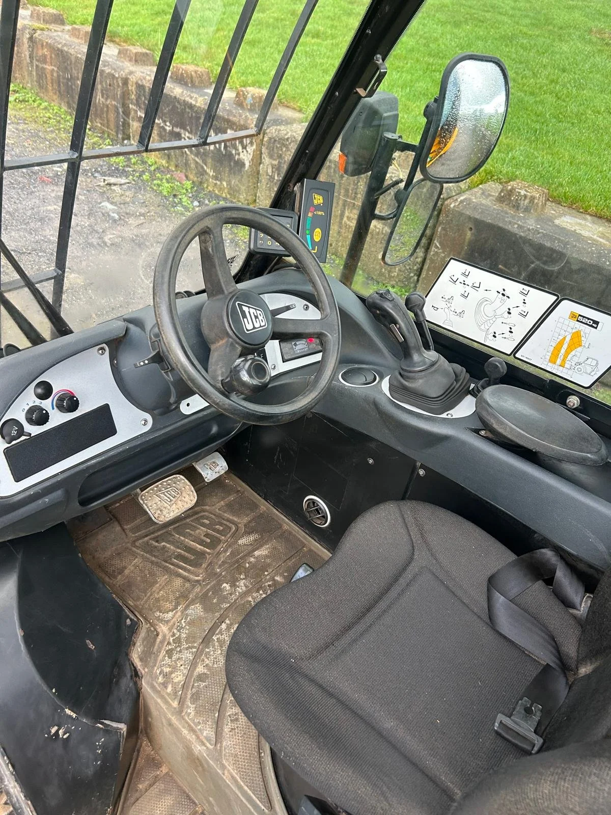 Interior view of a JCB construction vehicle, showing the steering wheel, control levers, dashboard, and a seat, with outside greenery visible through the window.
