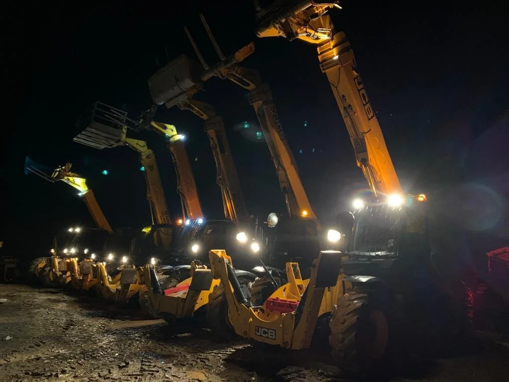Line of construction or excavation vehicles with all-terrain tires and boom arms extended, parked on dirt at night under dark sky with bright headlights illuminating the scene.