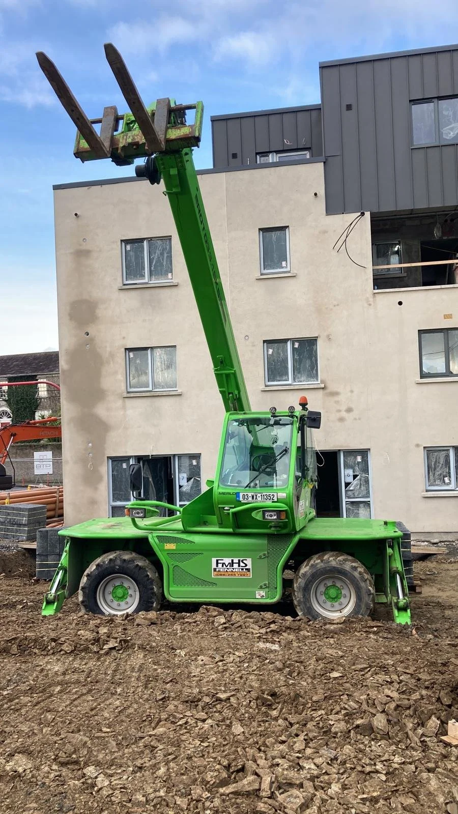 A green telescopic forklift with extended arm and prongs at a construction site in front of a multi-story building under construction.