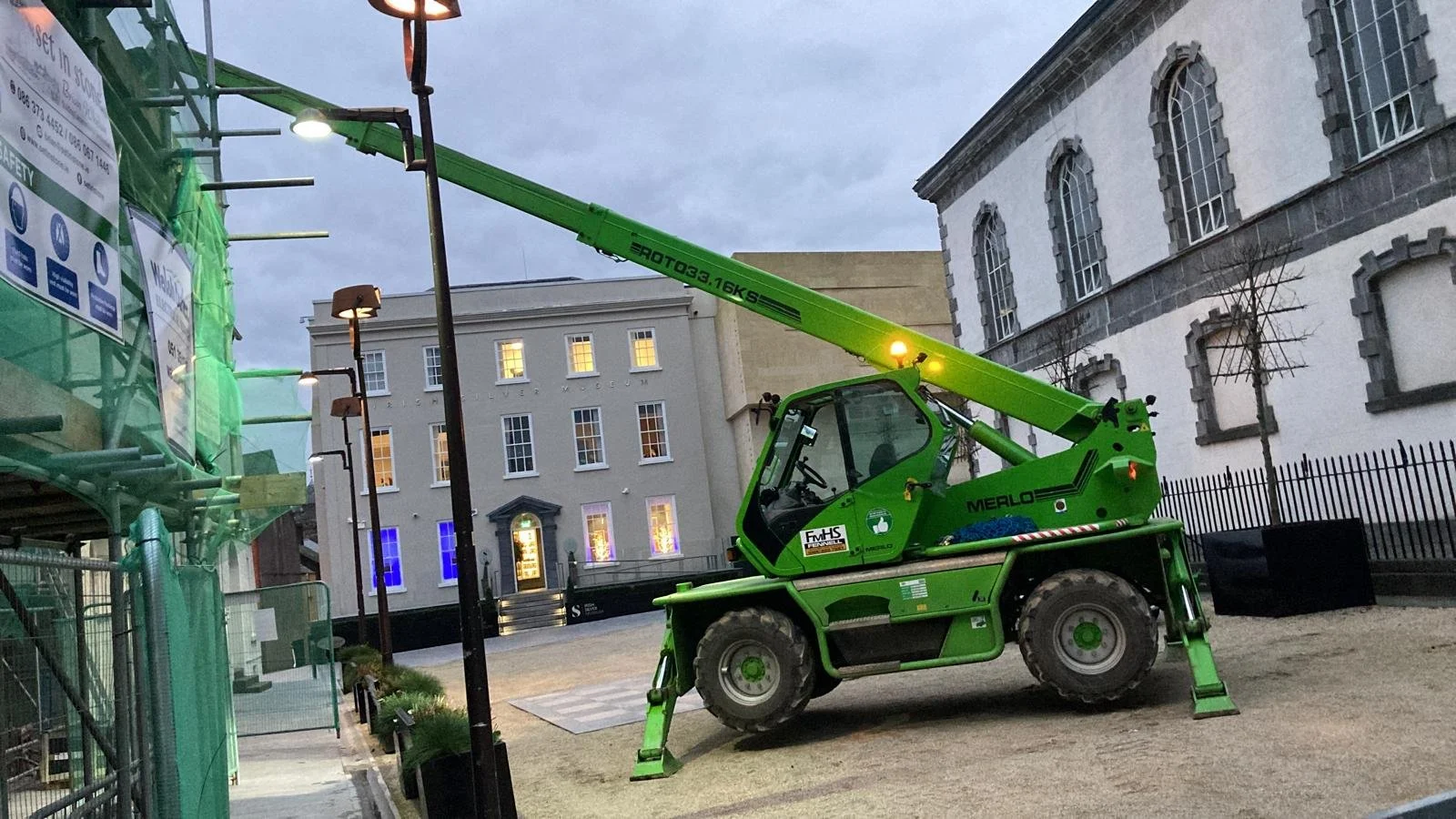 A bright green tower crane on wheels set up on a city street during evening hours, with a tall gray building in the background and a green construction site fence on the left.