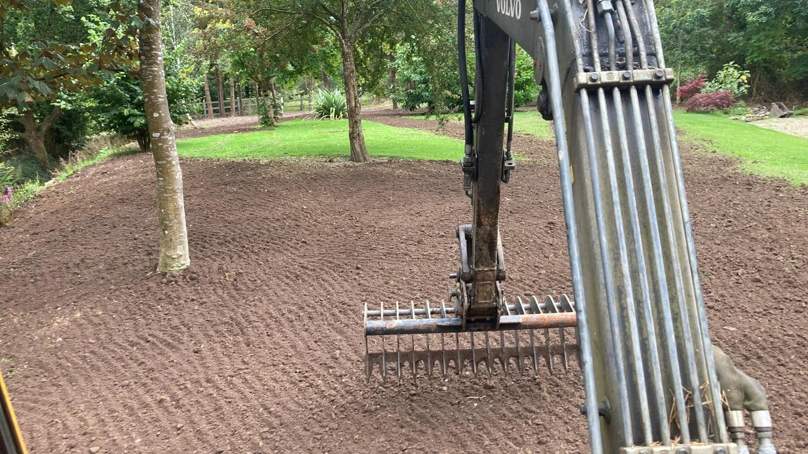 The image shows an excavator with its arm and bucket positioned near a freshly tilled dirt area in a backyard, surrounded by trees, grass, and landscaped plants.