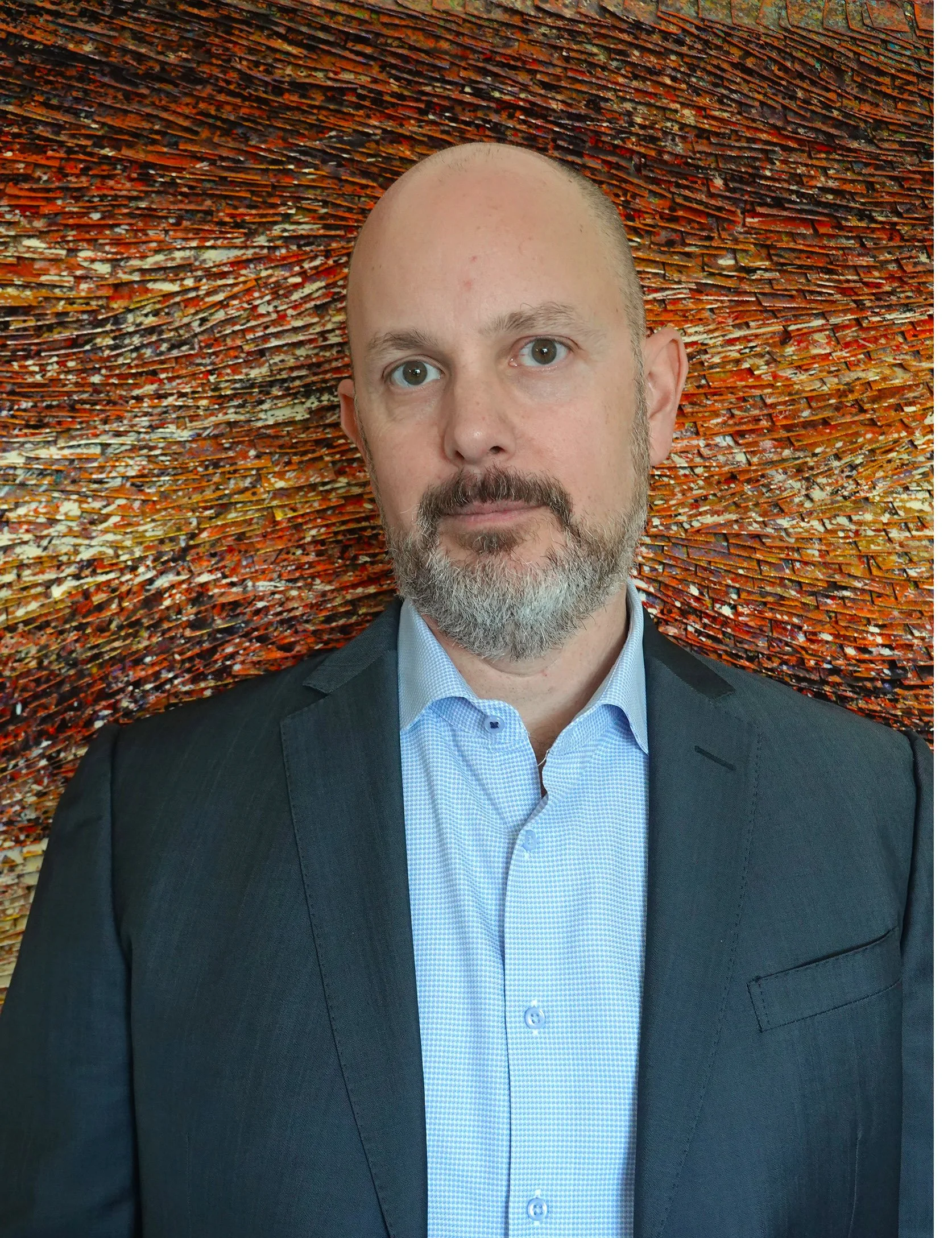 Portrait of a middle-aged man with a beard and mustache, wearing a dark blazer and light blue dress shirt, standing in front of an abstract textured background with orange and brown hues.