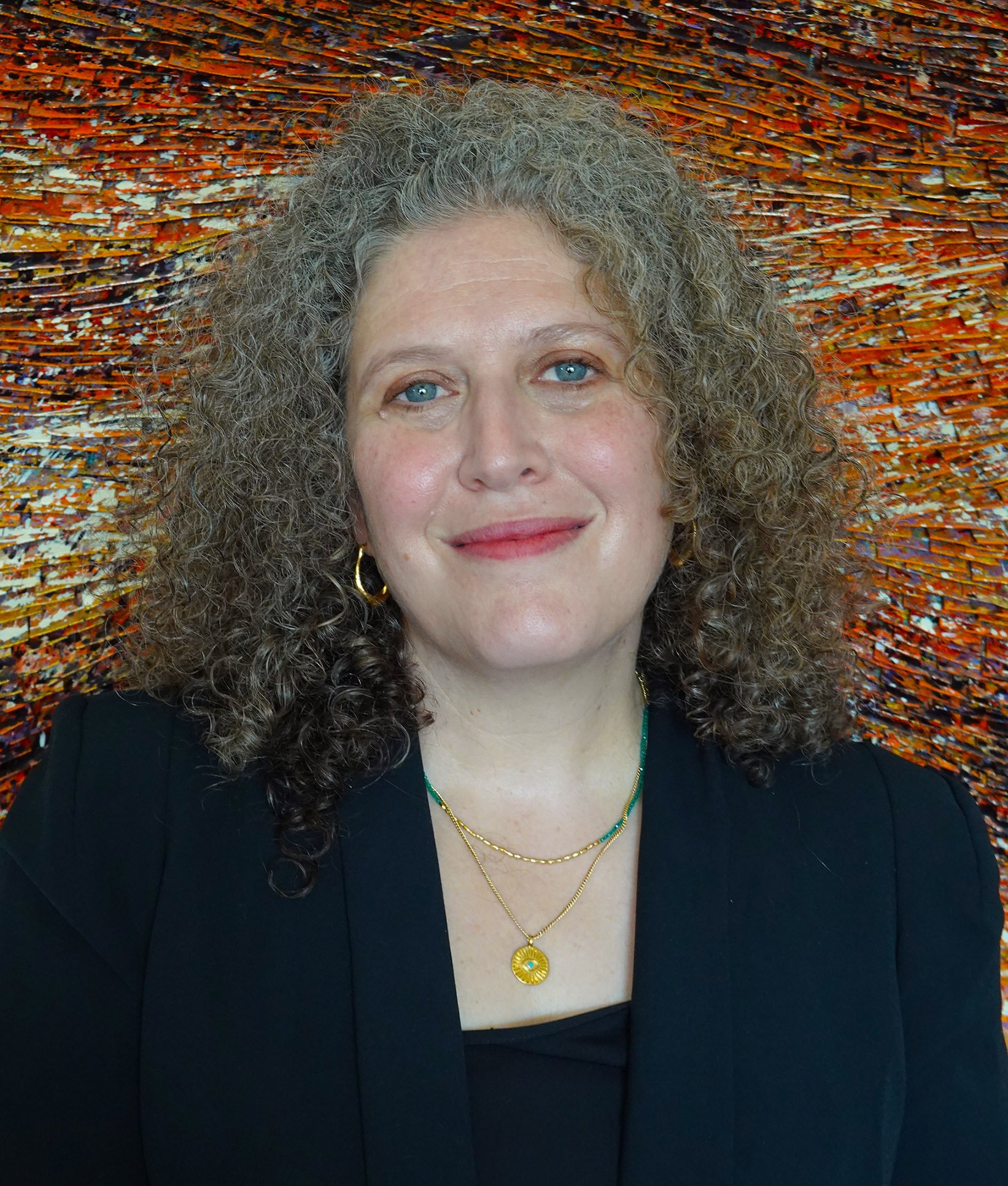 Lawyer Teri Stein with curly gray hair, blue eyes, and light skin, smiles at the camera, wearing gold jewelry and a black top or jacket. The background features an abstract, colorful, textured art piece with orange and reddish hues.