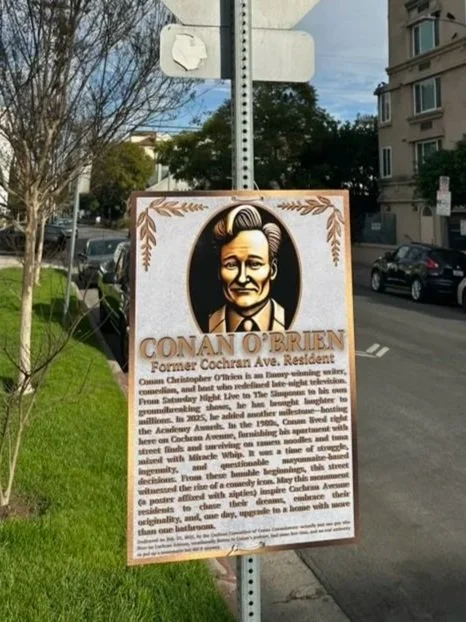 Historical plaque portrait of Conan O'Brien, former resident of Cochran Avenue, on a city sidewalk with cars and trees in the background.