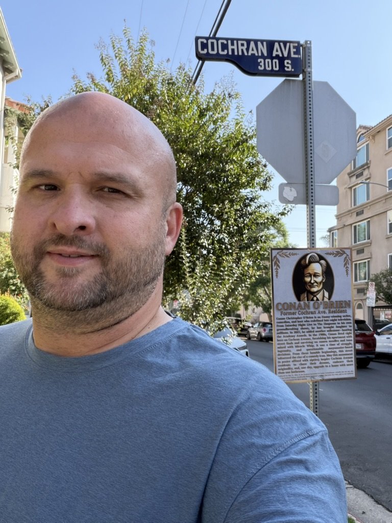 A man taking a selfie on Cochran Avenue in front of a street sign, with a historical plaque featuring a portrait of Conan O'Brien and a tree in the background.