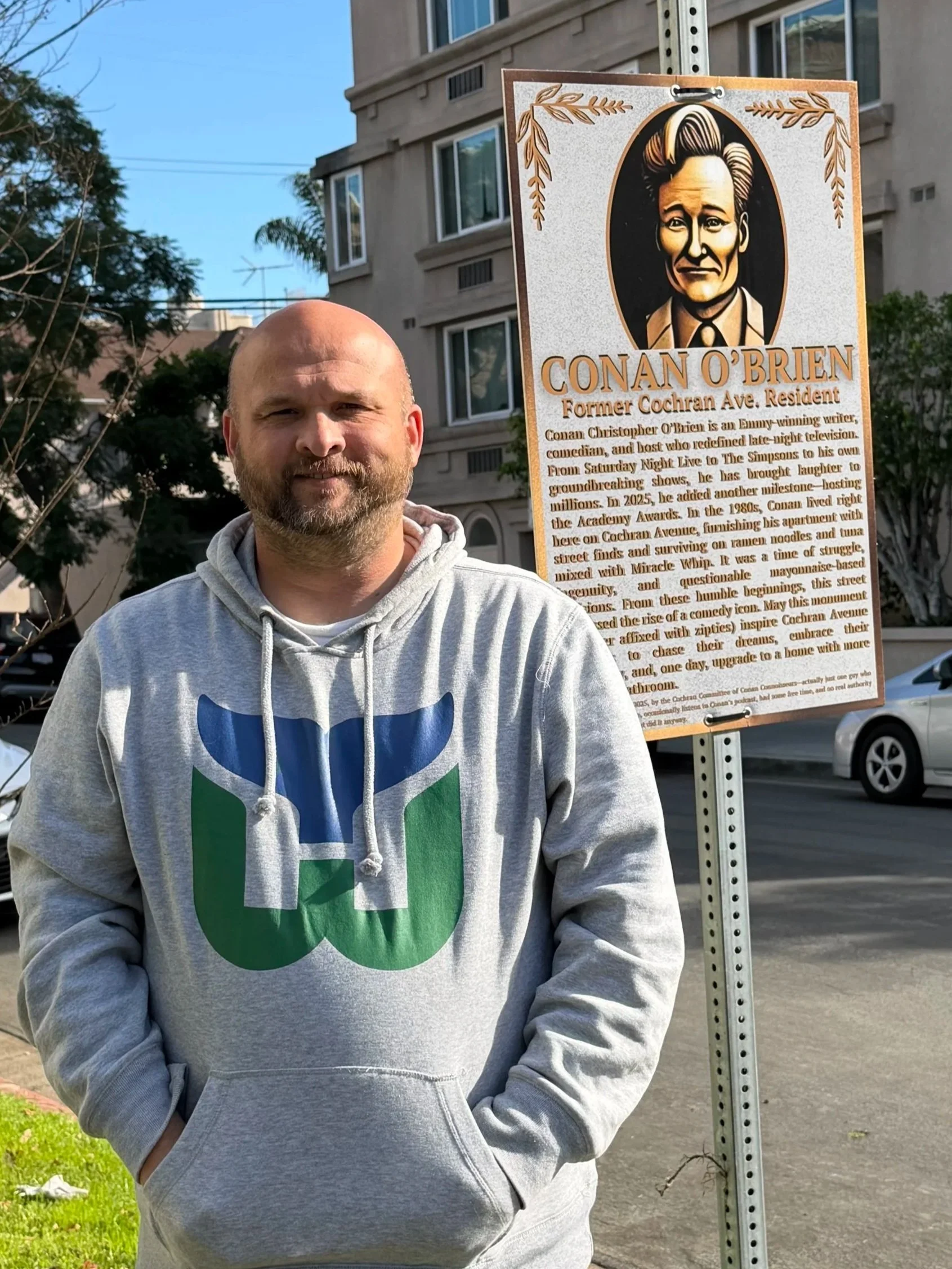 A man standing outdoors, wearing a gray hoodie with a blue and green logo, next to a sign honoring Conan O'Brien with biography details, on a city street with buildings and parked cars.
