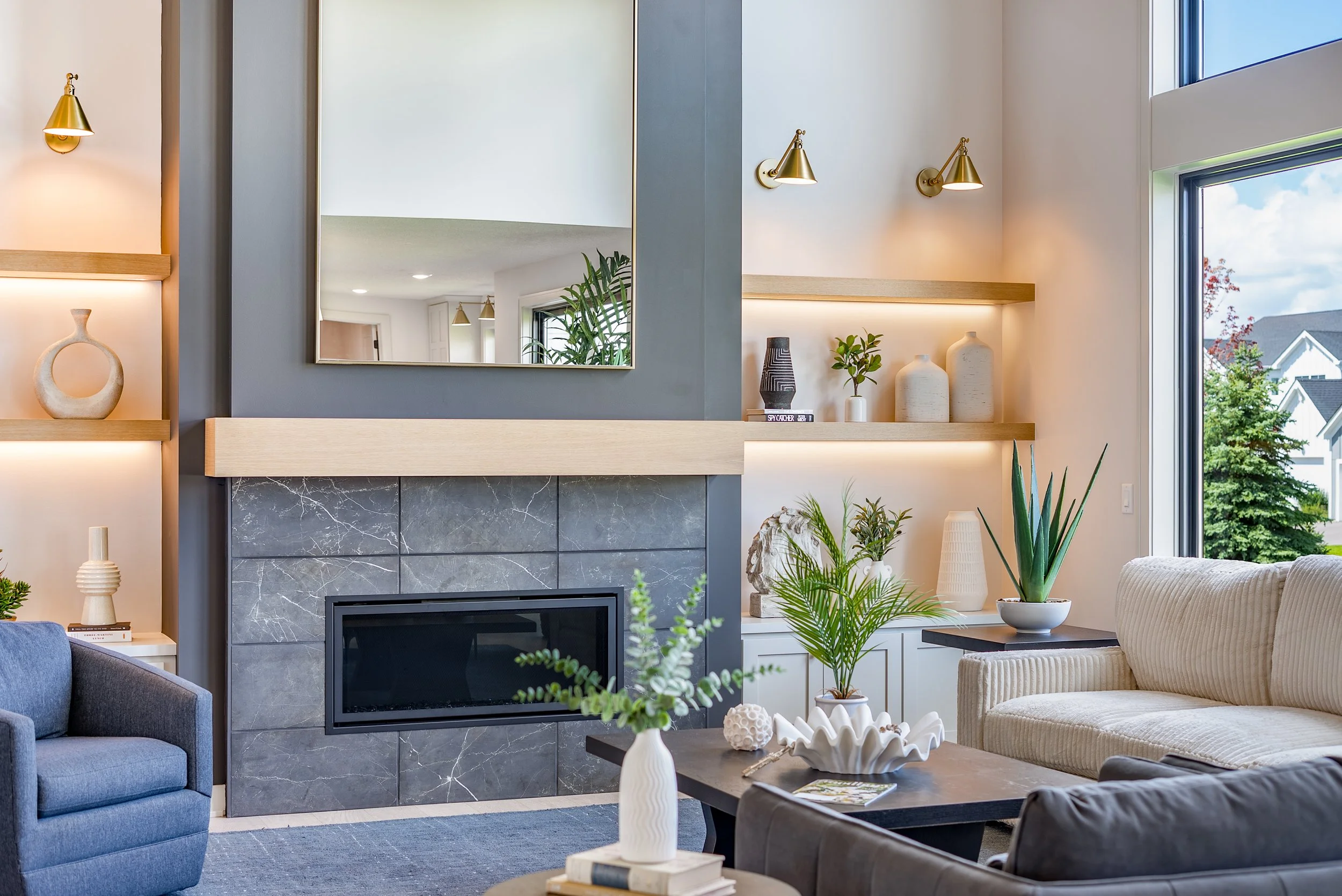 Living room with modern fireplace, gray and white decor, indoor plants, and large window showing outdoors with trees.