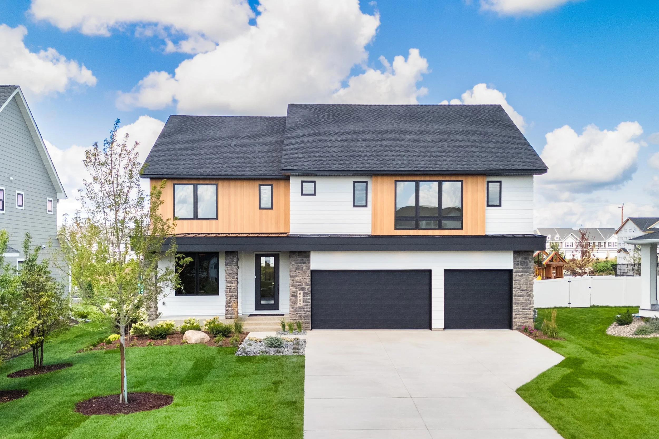 Modern two-story house with a black roof, mix of white and wooden siding, stone accents, black garage doors, and a well-maintained lawn with trees and landscaping.