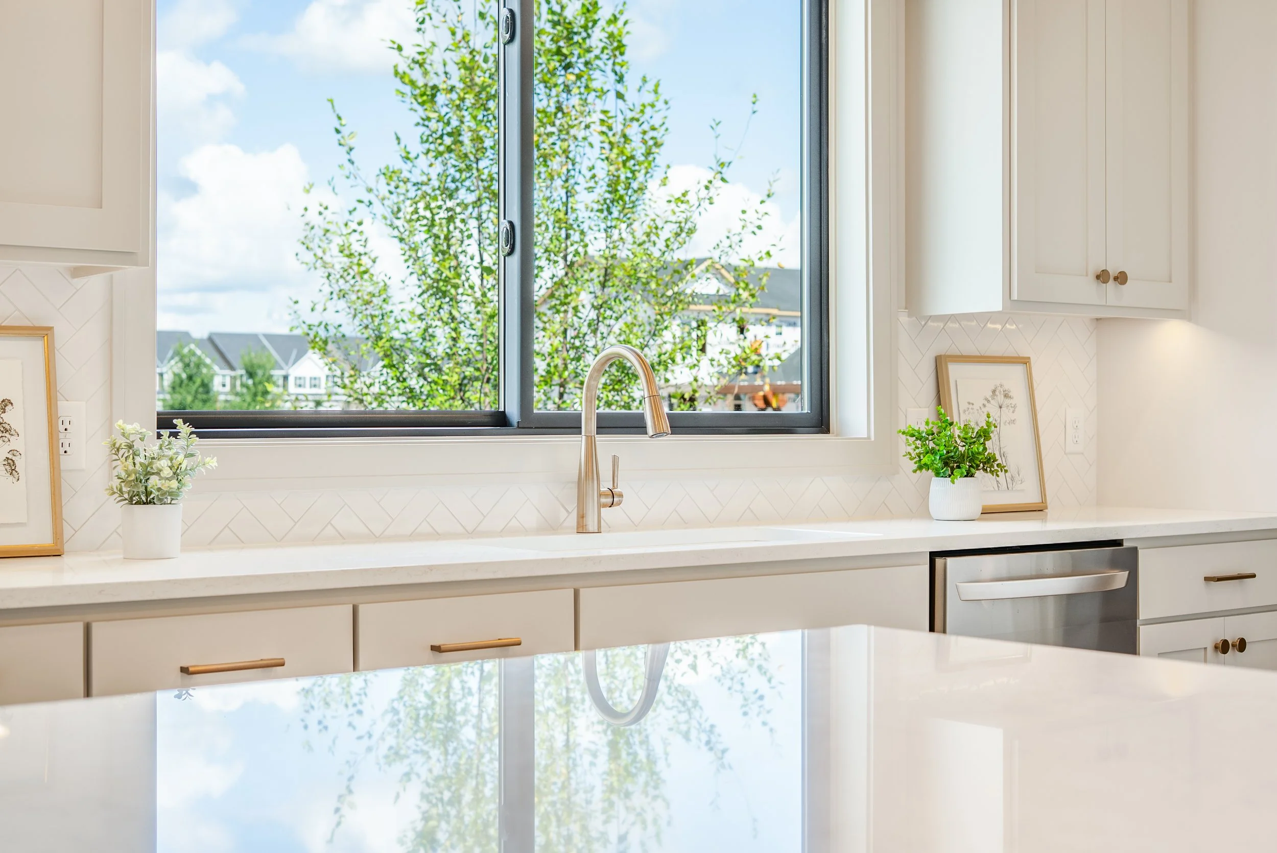 A modern kitchen with a large window showing a view of trees and houses outside. The kitchen features a white countertop, cabinetry, a gold faucet, and potted plants for decoration.