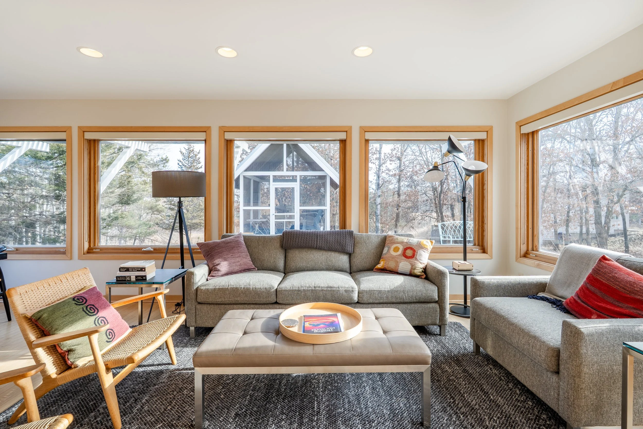 Living room with large windows, beige sofas, colorful pillows, a tufted ottoman with a tray, wooden armchair with a patterned pillow, and floor lamps.