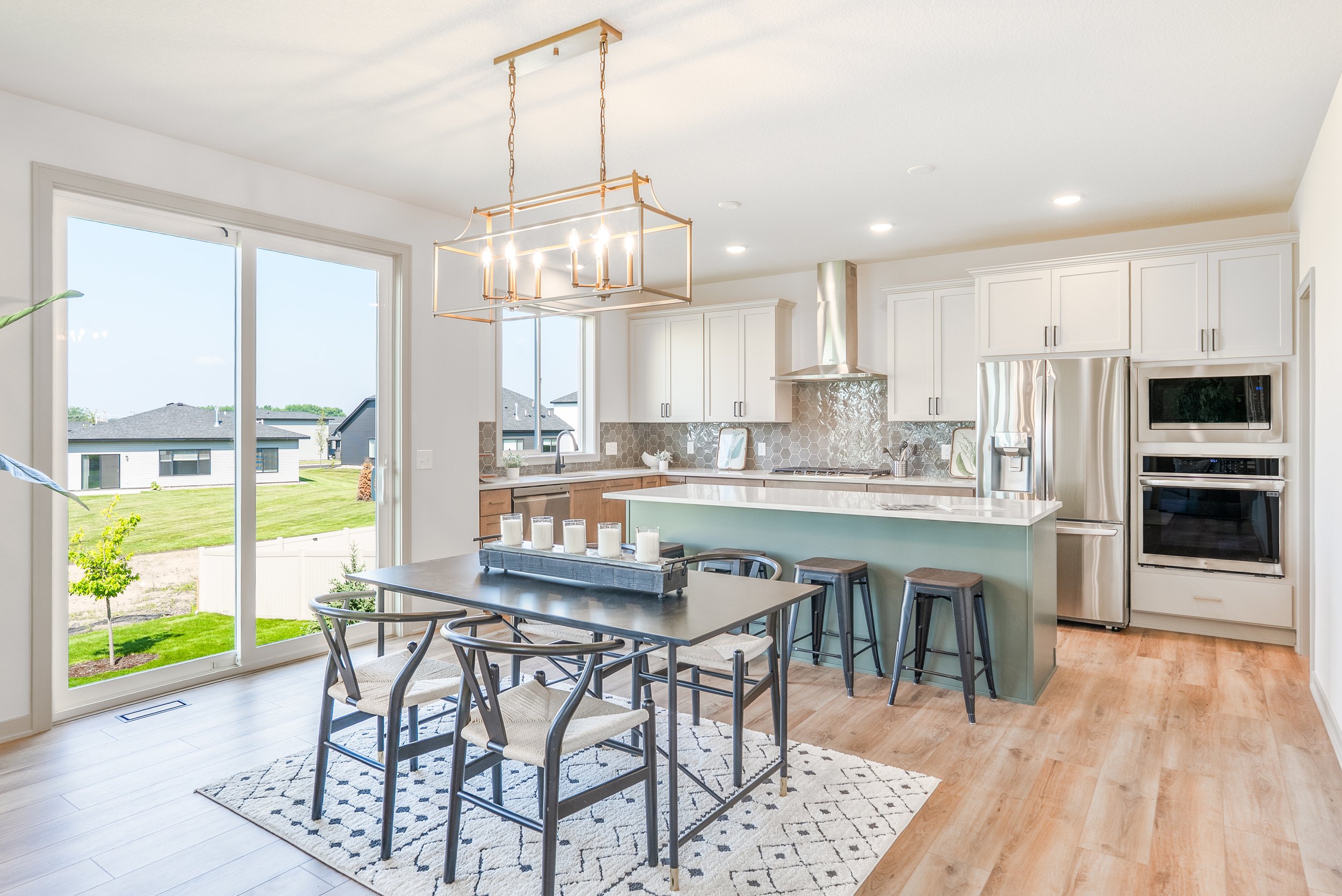 Bright modern kitchen with white cabinets, stainless steel appliances, and a central island with bar stools. A dining table with chairs is in front of a large sliding glass door leading outside.