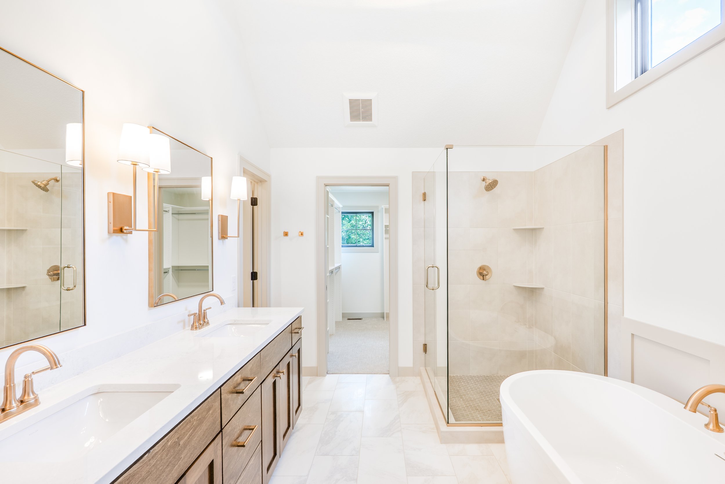 Bright modern bathroom with double vanity sinks, a large mirror, gold fixtures, a glass-enclosed shower, a freestanding bathtub, and a small window.