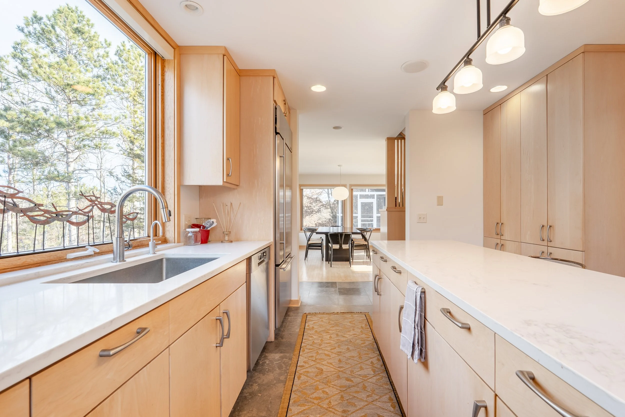 Kitchen with light wood cabinets, white marble countertops, stainless steel appliances, large window overlooking trees, dining area with round table and black chairs, ceiling lights, beige walls, and a narrow rug on wood and tile floor
