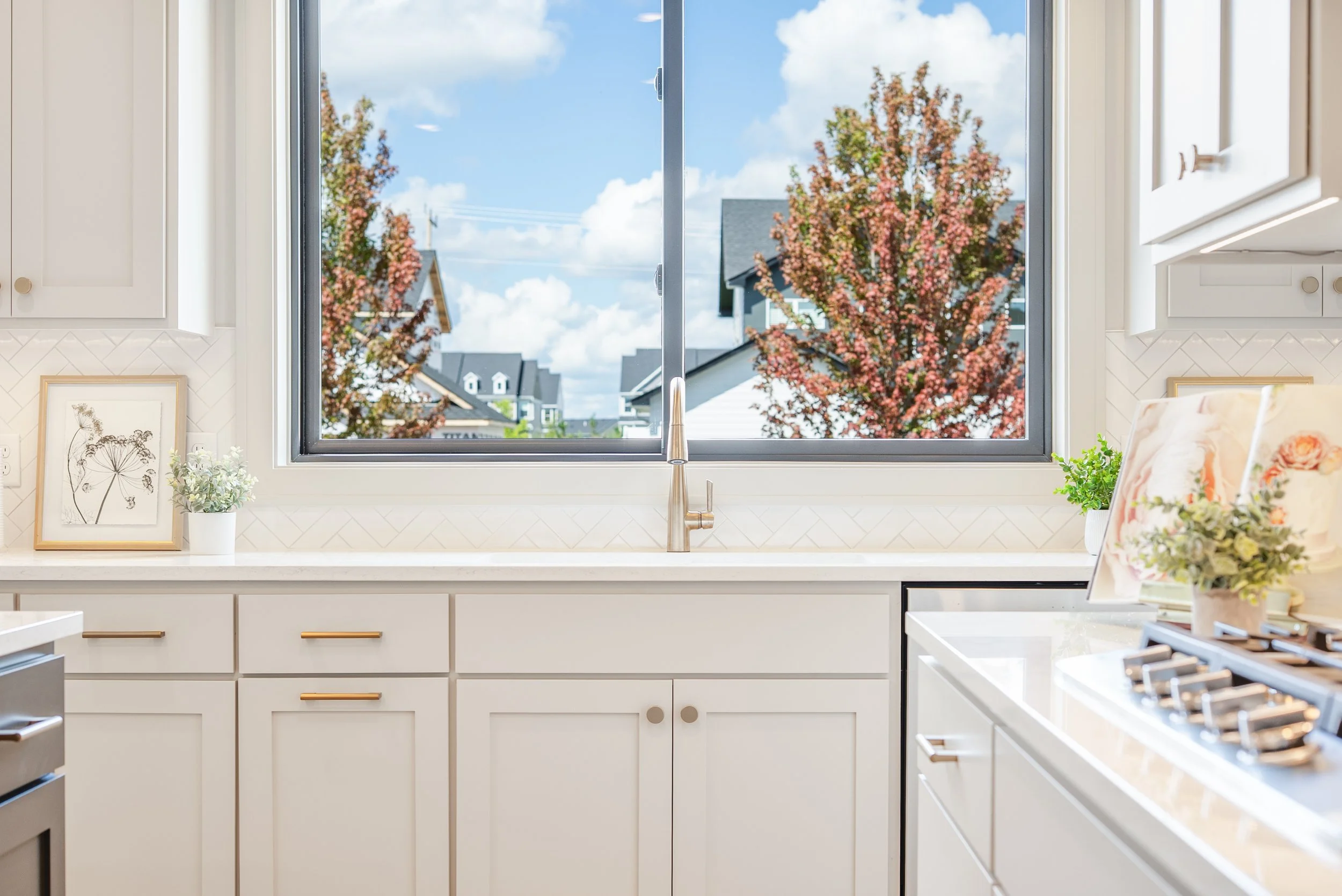 Bright kitchen with a large window overlooking trees and houses, white cabinets, a framed botanical print and potted plants on the countertop.