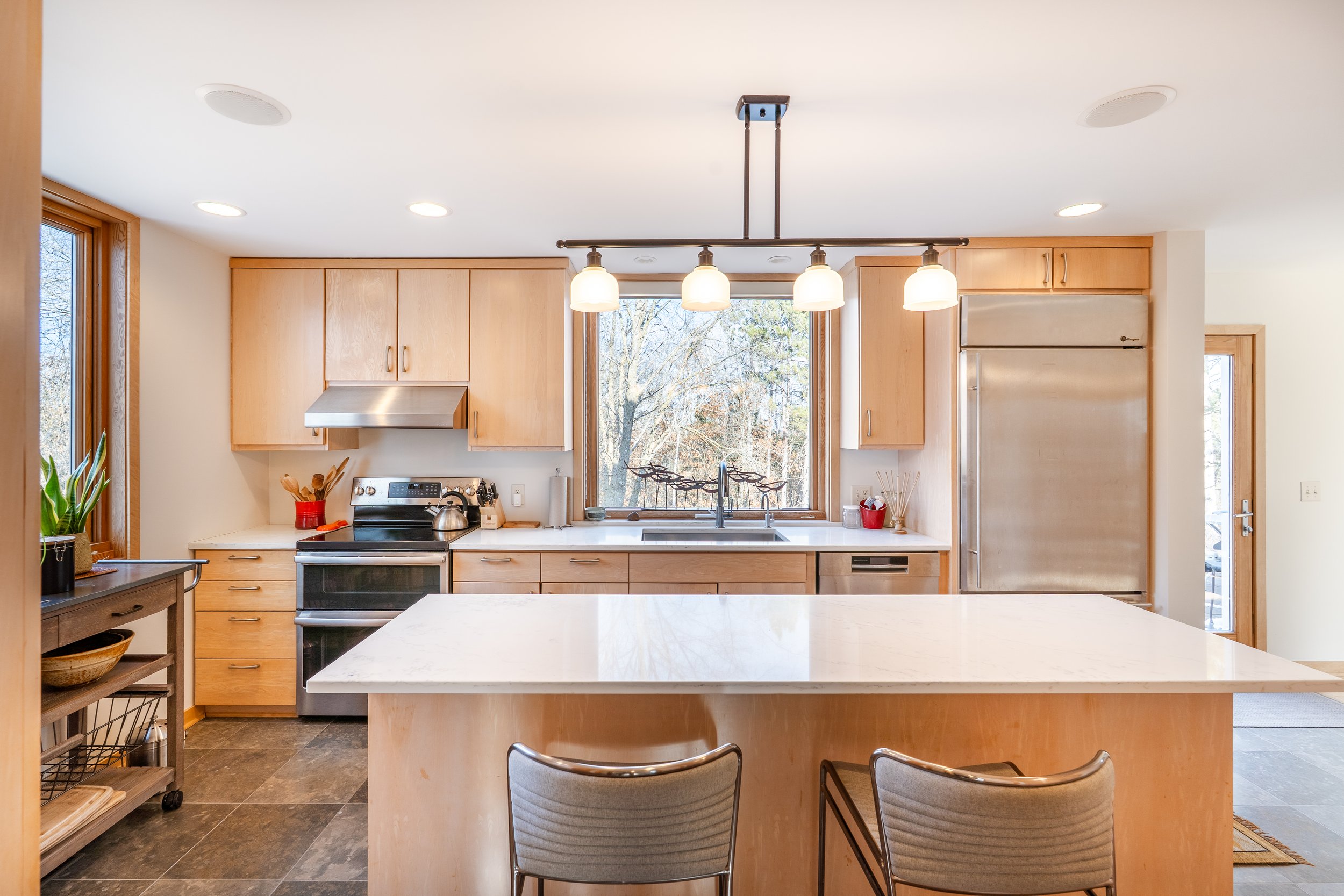 Modern kitchen with light wood cabinets, stainless steel appliances, and a large island with a white marble countertop. There is a window over the sink and a hanging light fixture with four bulbs.
