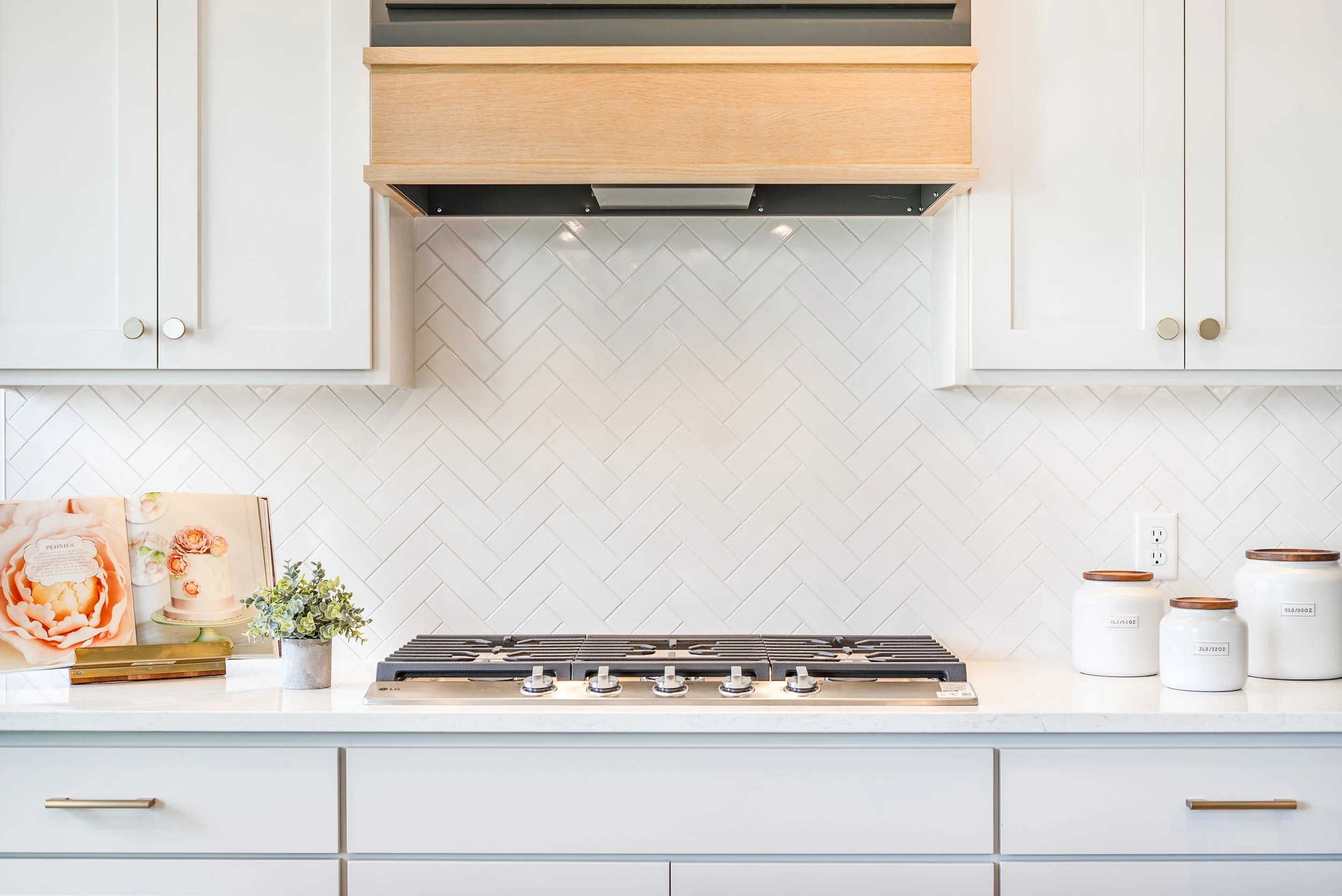Modern white kitchen with a gas stovetop, decorative jars, and a small potted plant on the countertop. White cabinets with round knobs and a white, textured tile backsplash.