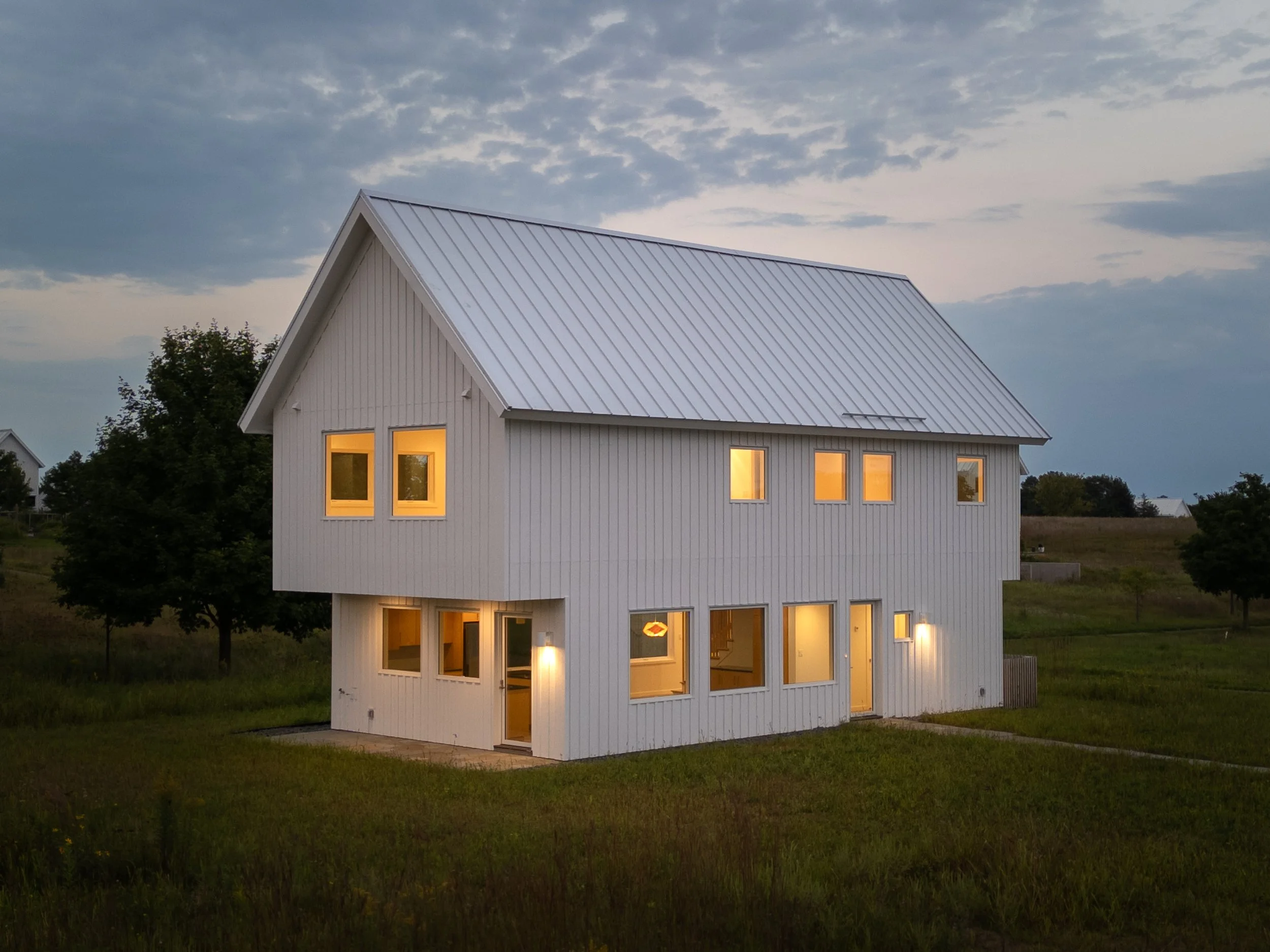 A modern two-story house with white siding and a metal roof, illuminated from inside, in a grassy open area during dusk with a partly cloudy sky.