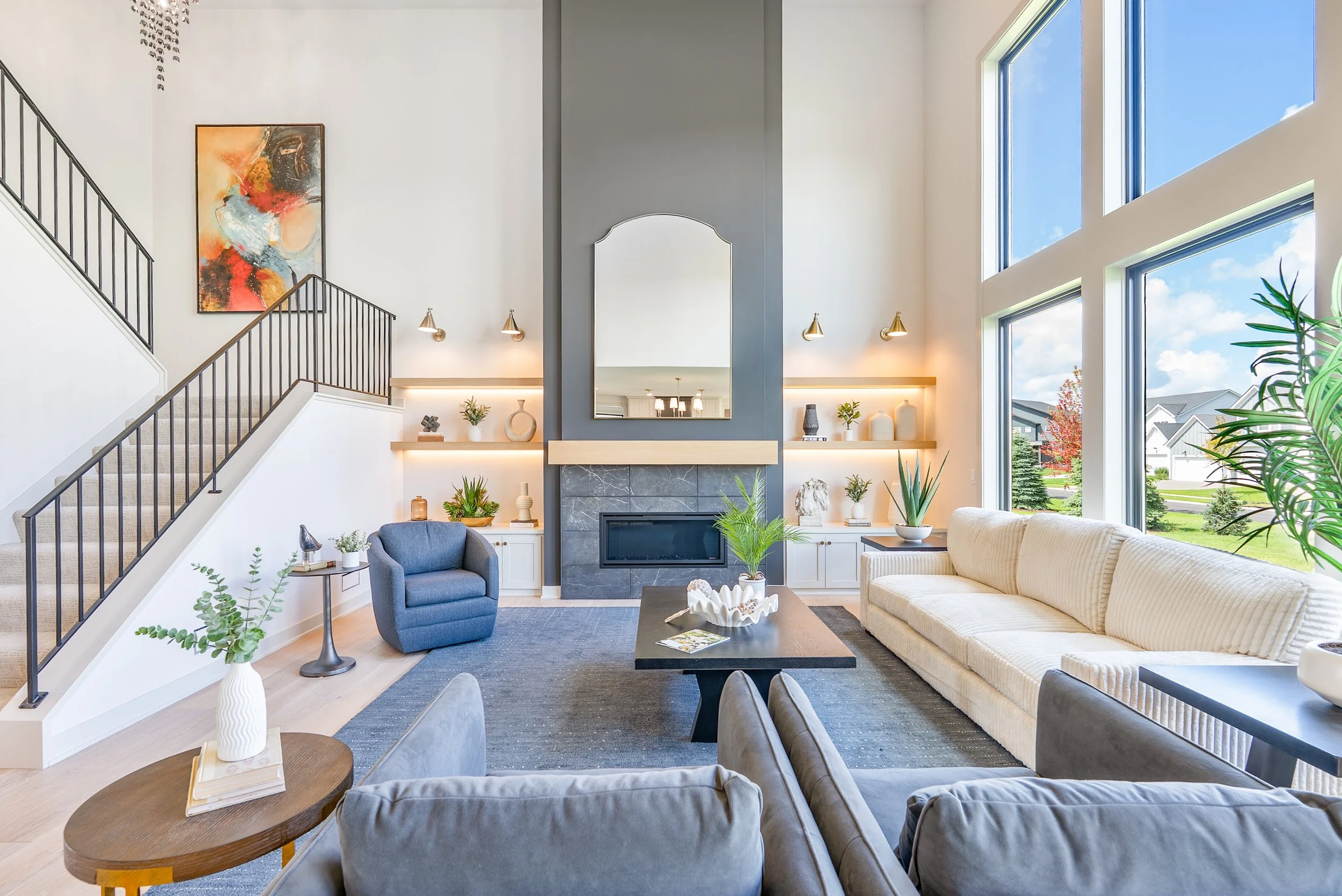 Bright living room with large windows, white sofa, blue armchair, coffee table, and decorative shelves with plants, vases, and artwork.