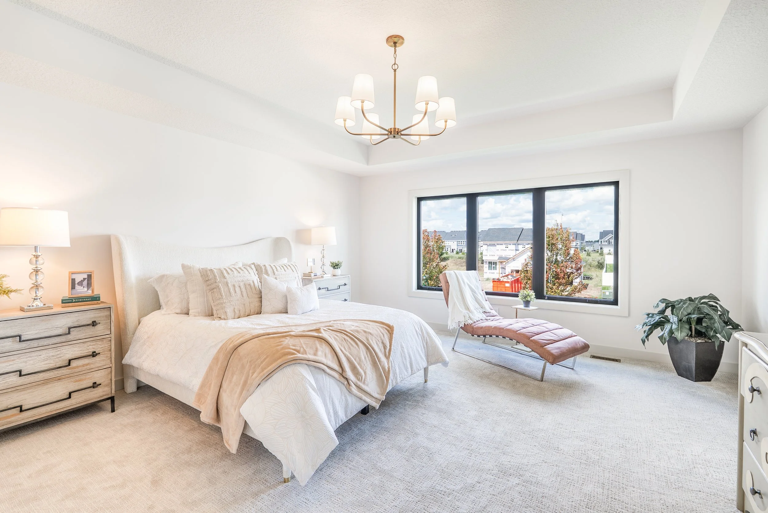 Bright bedroom with a large bed, bedside tables, lamps, and a window with a view of buildings and trees outside.
