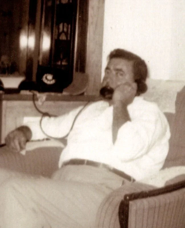 A man with dark hair and a beard, wearing a white shirt, lying on a bed and speaking on a corded phone, in a room with a cabinet and shelves behind him.