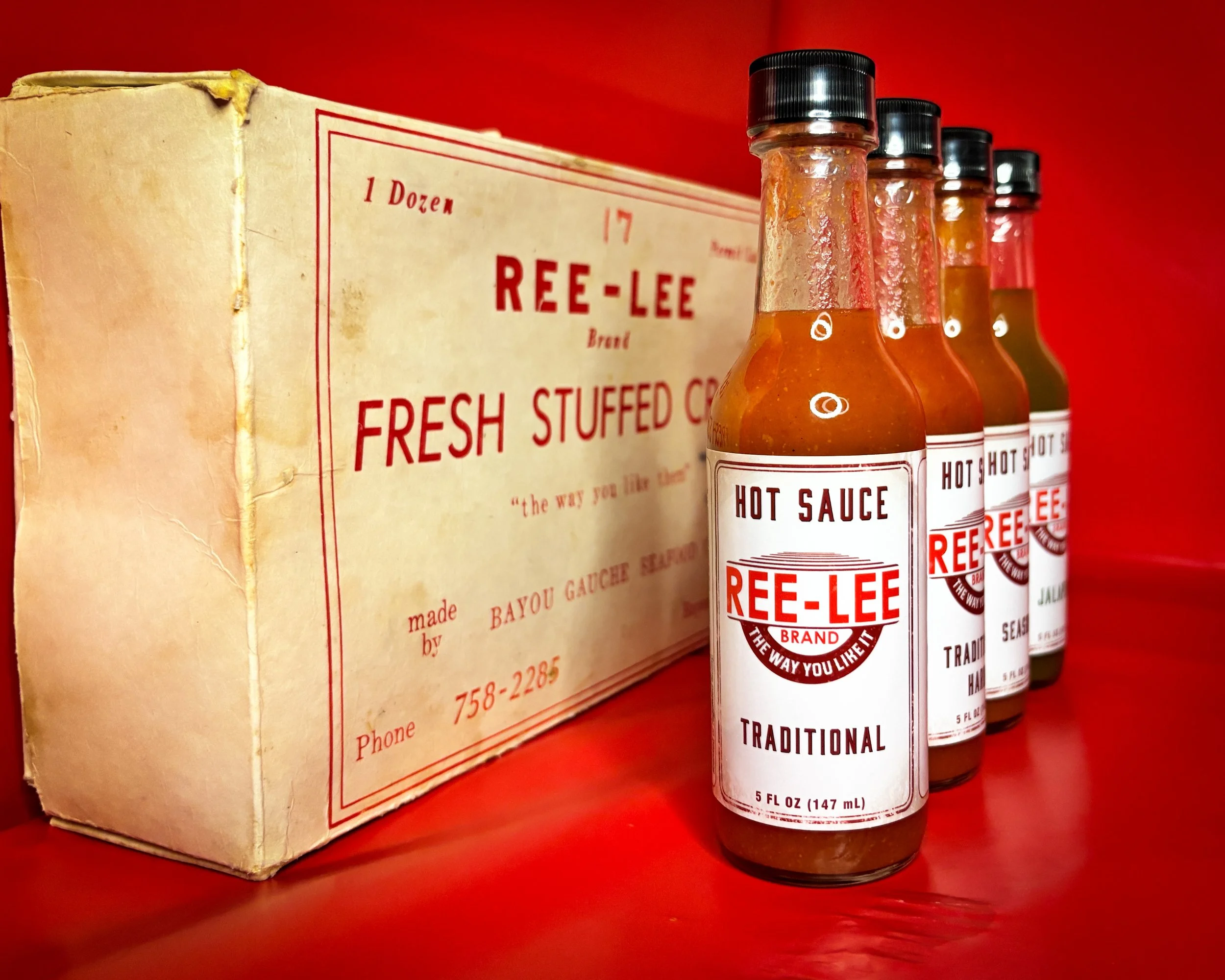 A row of Four bottles of Ree-Lee hot sauce standing in front of a boxed dozen of Ree-Lee brand hot sauce, on a red surface with a red background.