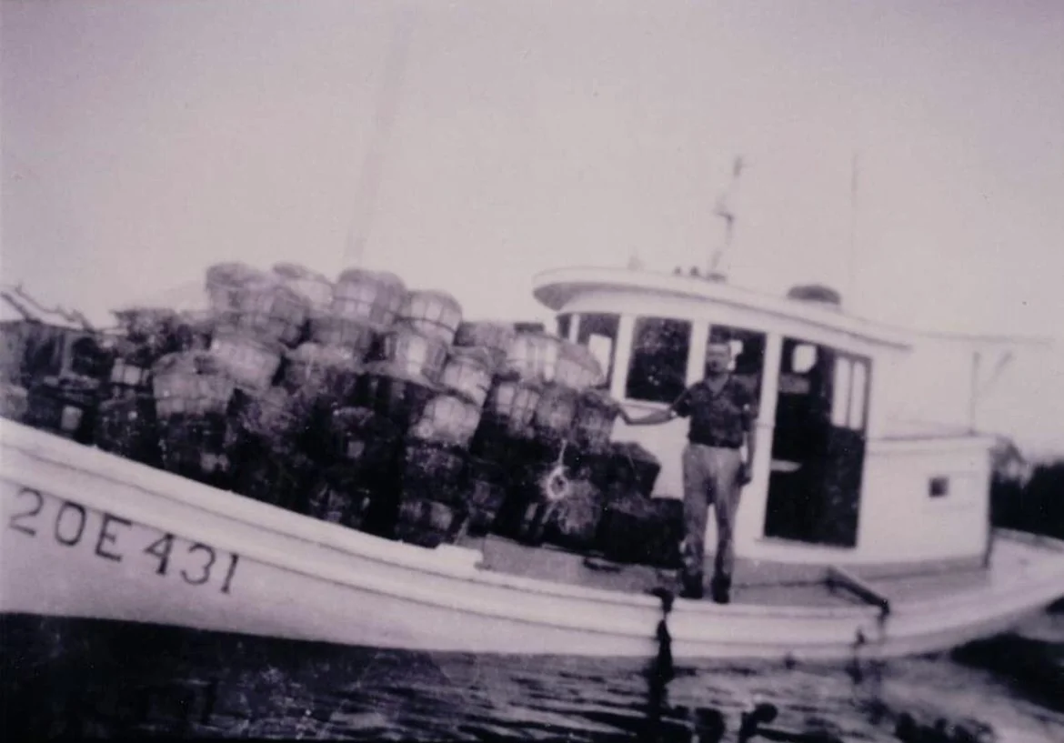 A person standing on a boat filled with stacked bottles, likely crates or containers, with water beneath.