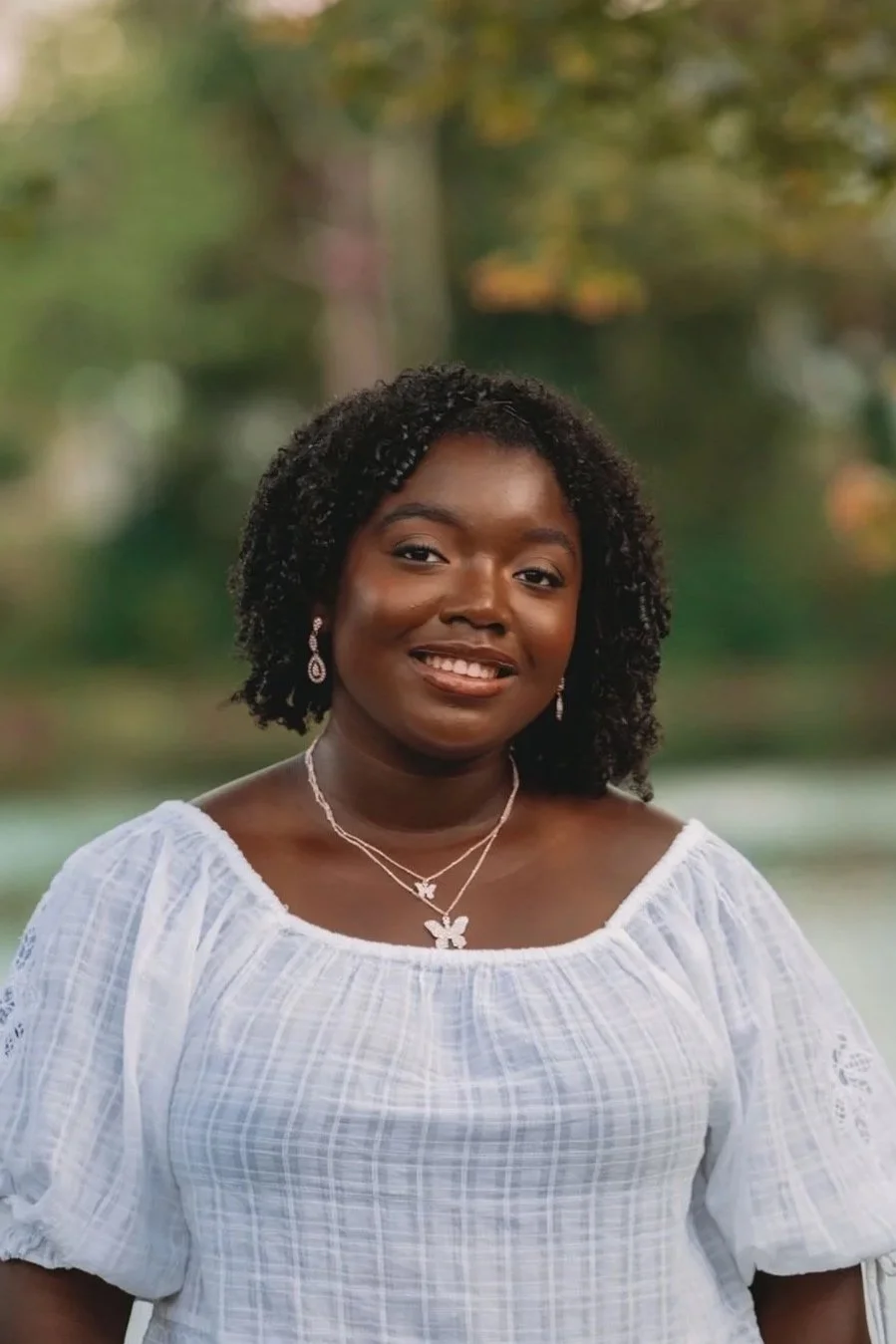 A woman with dark curly hair wearing a white blouse and jewelry, standing outdoors with a blurred green and autumnal background.