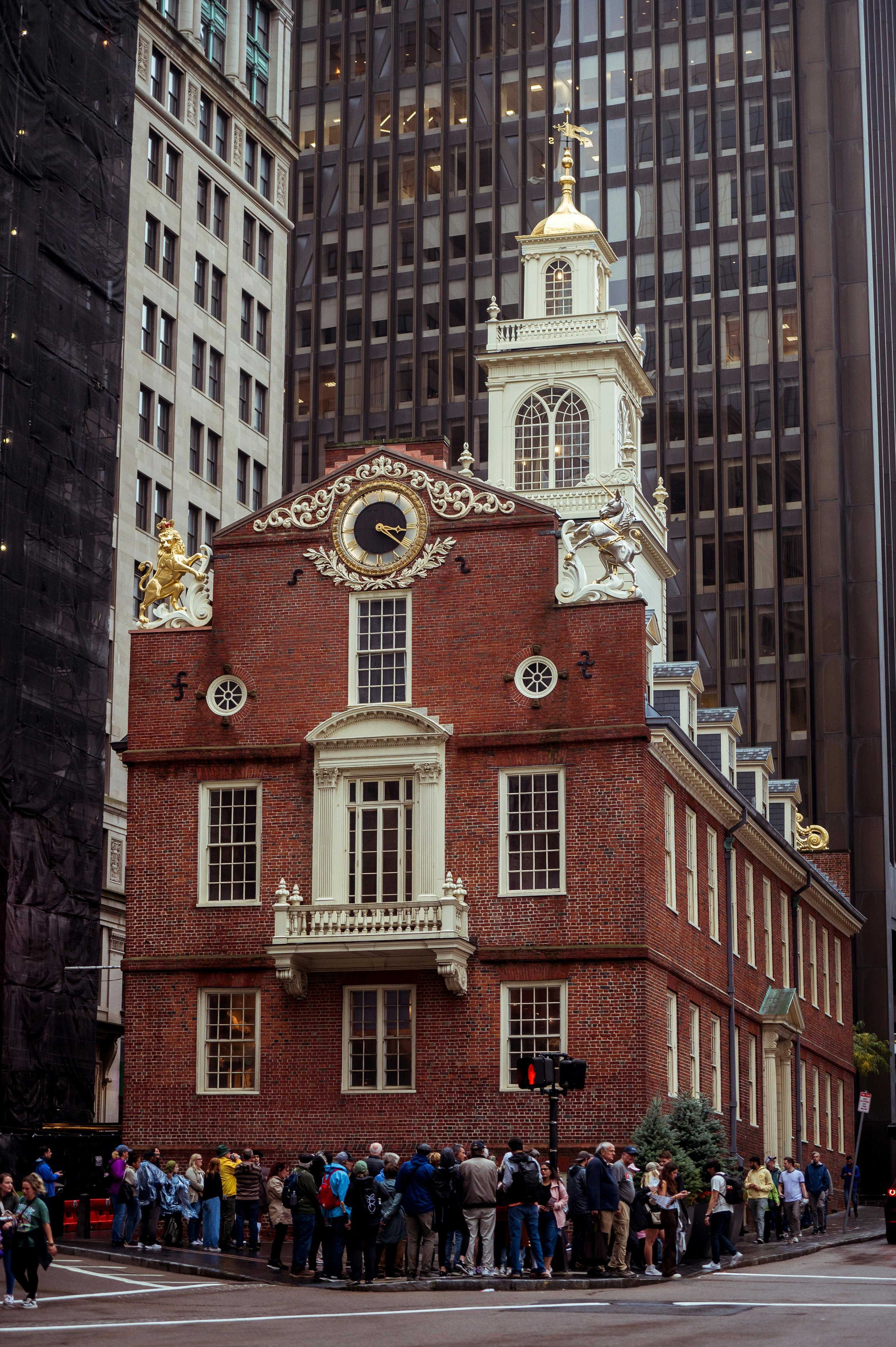 A historic red brick building with white trim and a clock, topped with a white tower and gold weather vane, standing among modern skyscrapers. A large group of people lines up outside on the street corner.