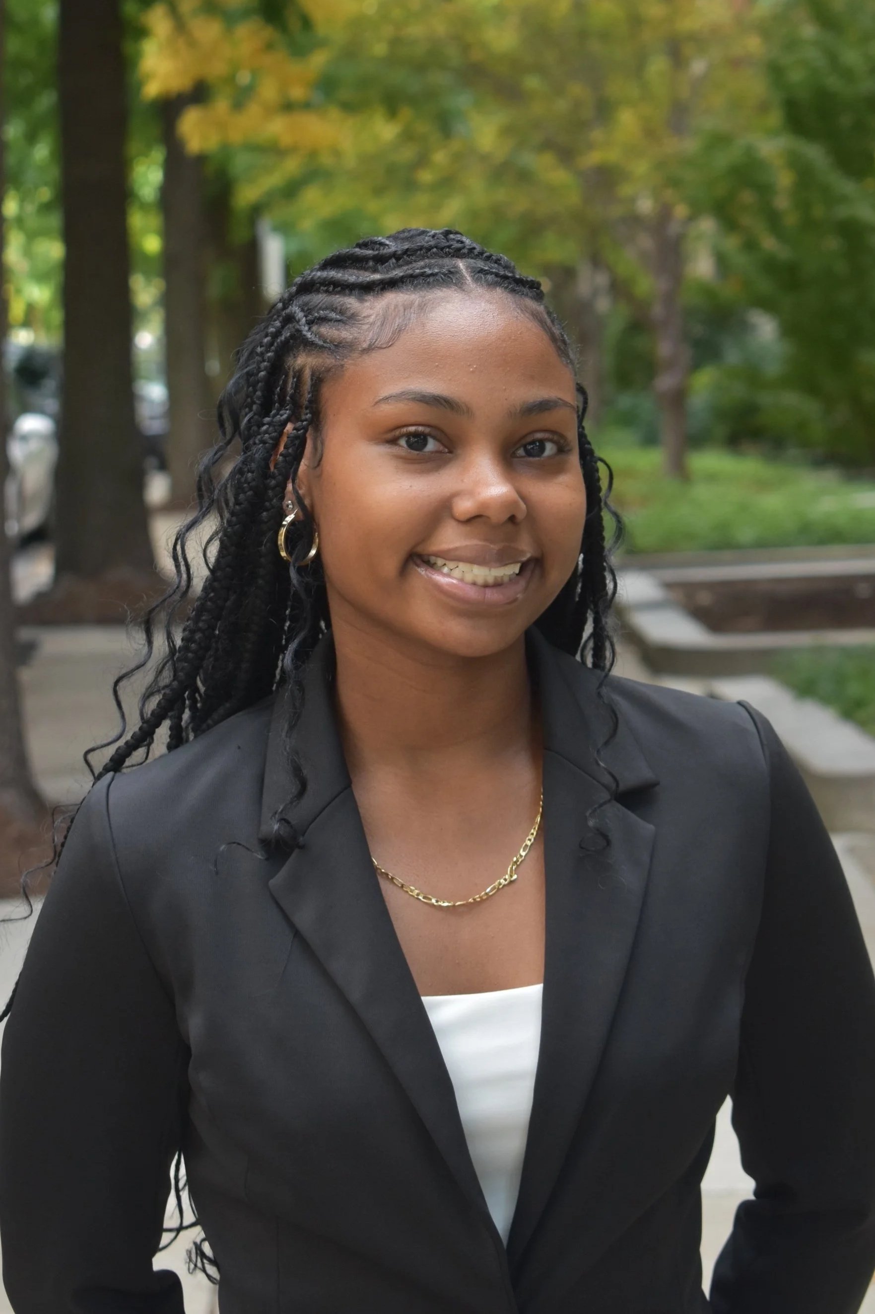 A young woman with long black curly braids, wearing a black blazer, white top, gold jewelry, smiling outdoors with trees and park benches in the background.