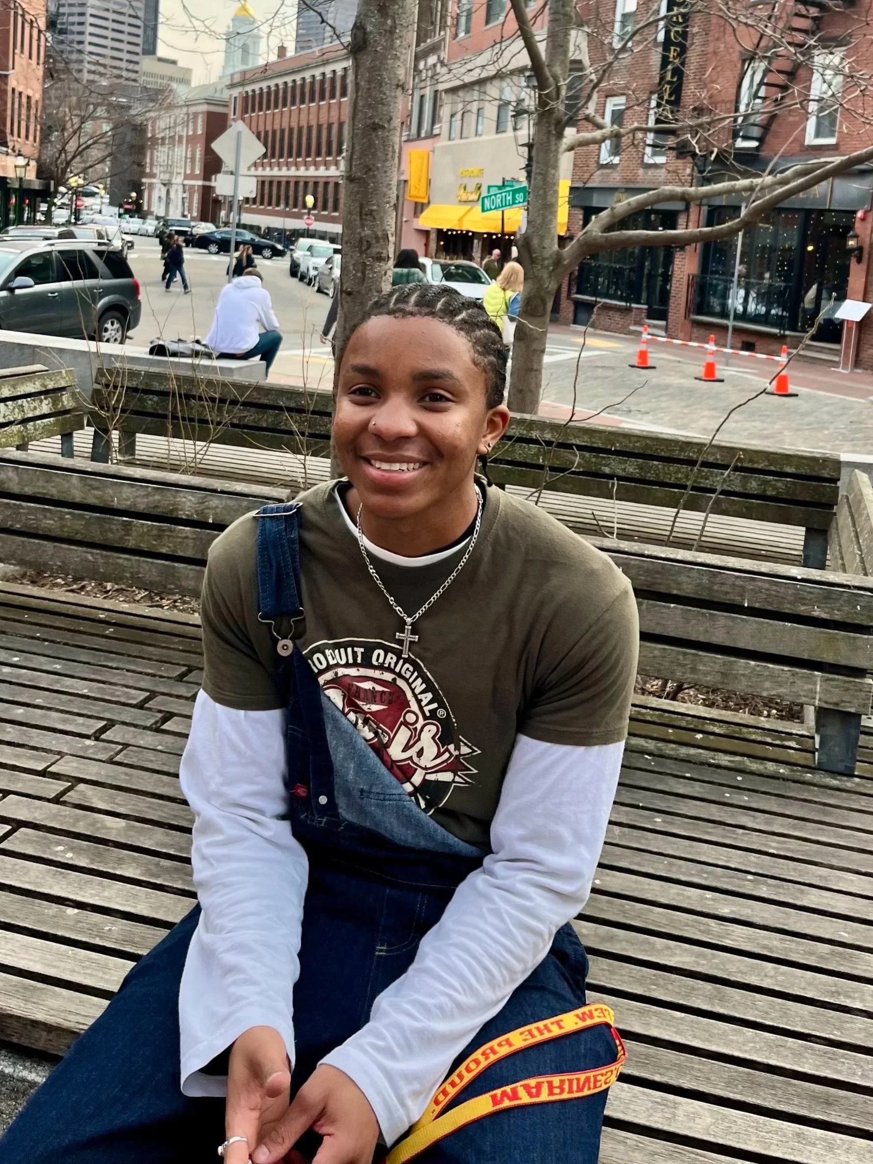 A young person with braids, smiling, sitting on a wooden bench on a city street with buildings, cars, and pedestrians in the background.
