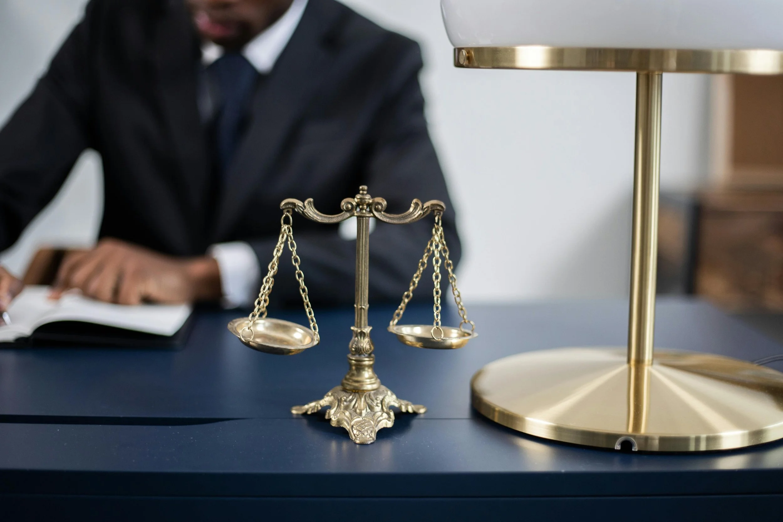 A golden scale of justice on a dark blue table, with a person in a suit writing in a notebook in the background.