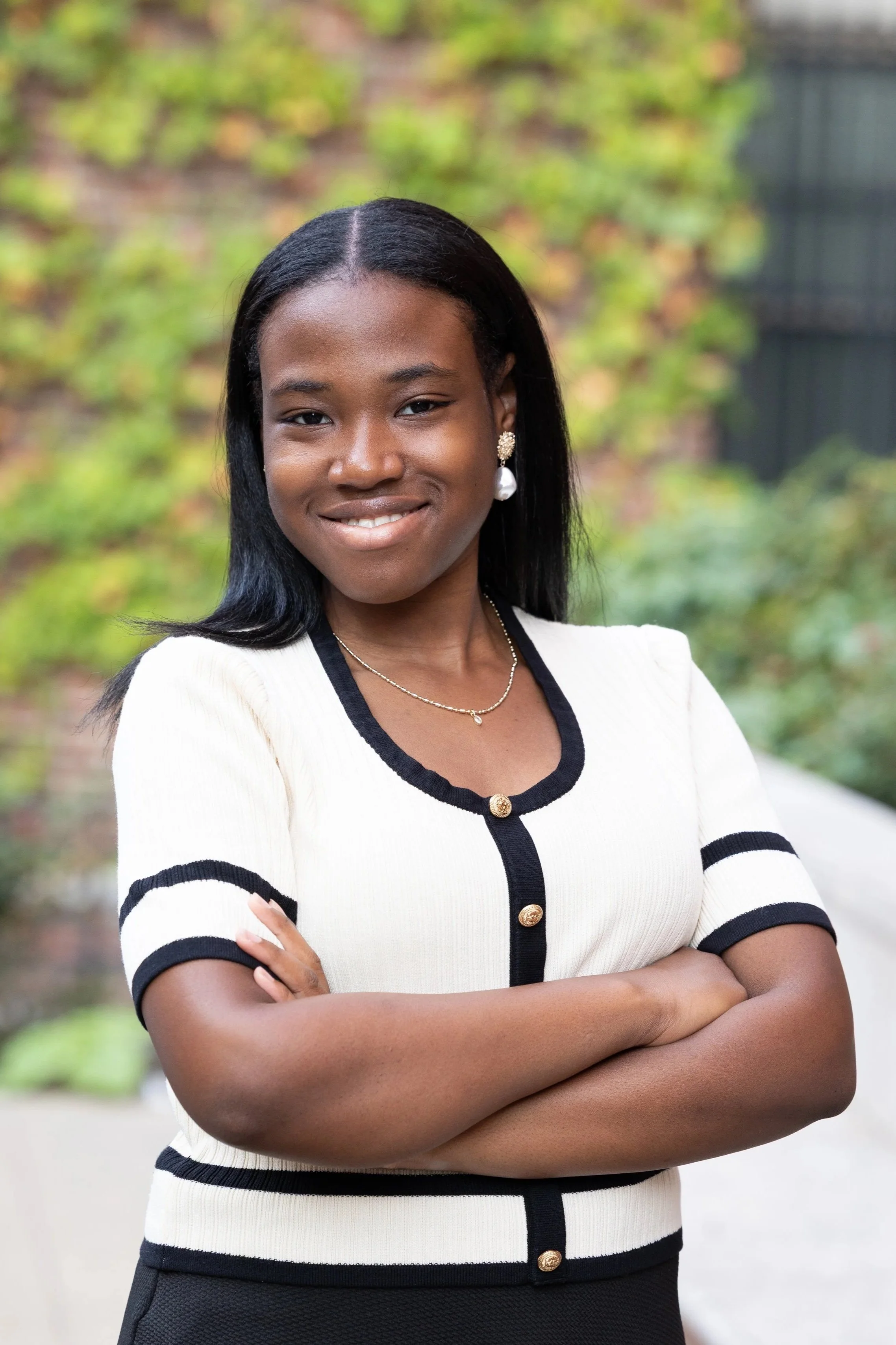 A portrait of a young Black woman standing outdoors with crossed arms, wearing a cream-colored short-sleeve top with black accents, pearl earrings, and a delicate necklace, smiling at the camera, with a blurred green and brick background.