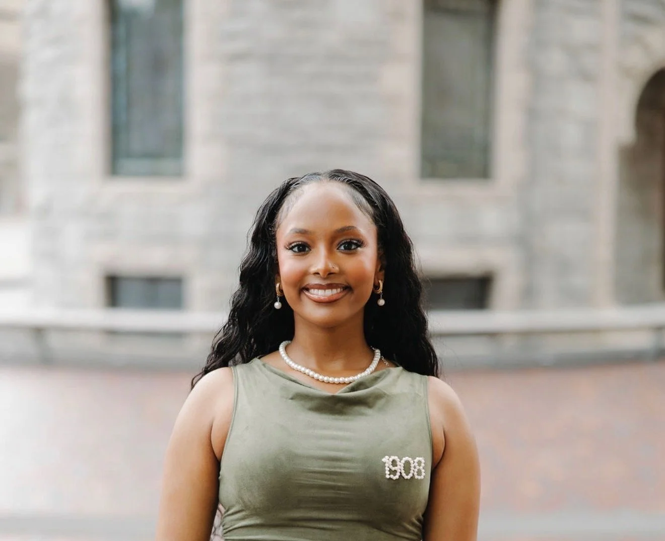 A young woman smiling outdoors, wearing a green sleeveless top with '1908' written on it, pearl earrings, and a pearl necklace.