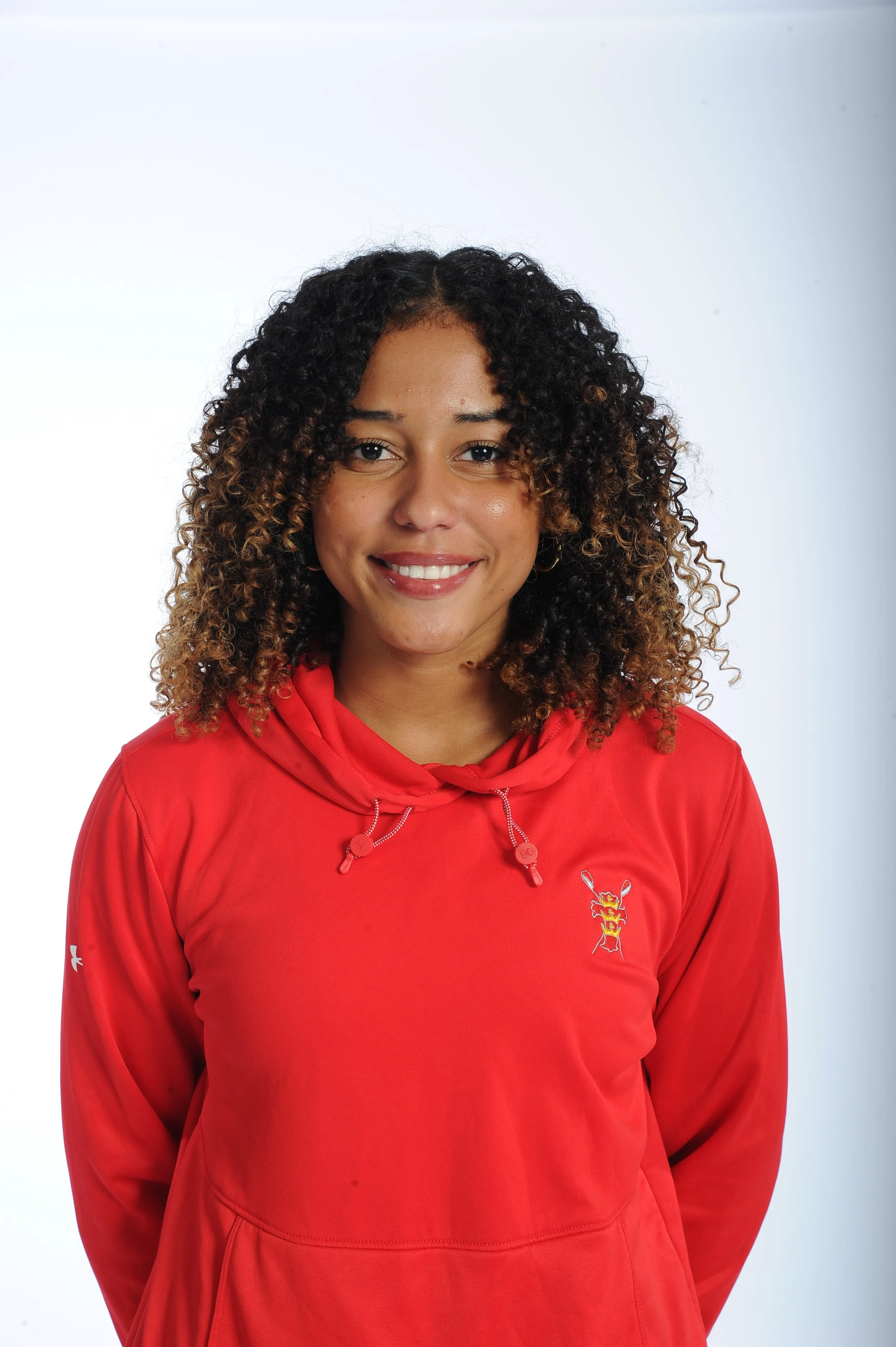 A woman with curly hair wearing a red athletic hoodie smiling at the camera against a white background.