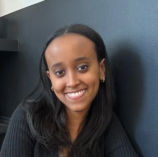 A young woman with dark hair and earrings smiling at the camera against a dark blue wall.