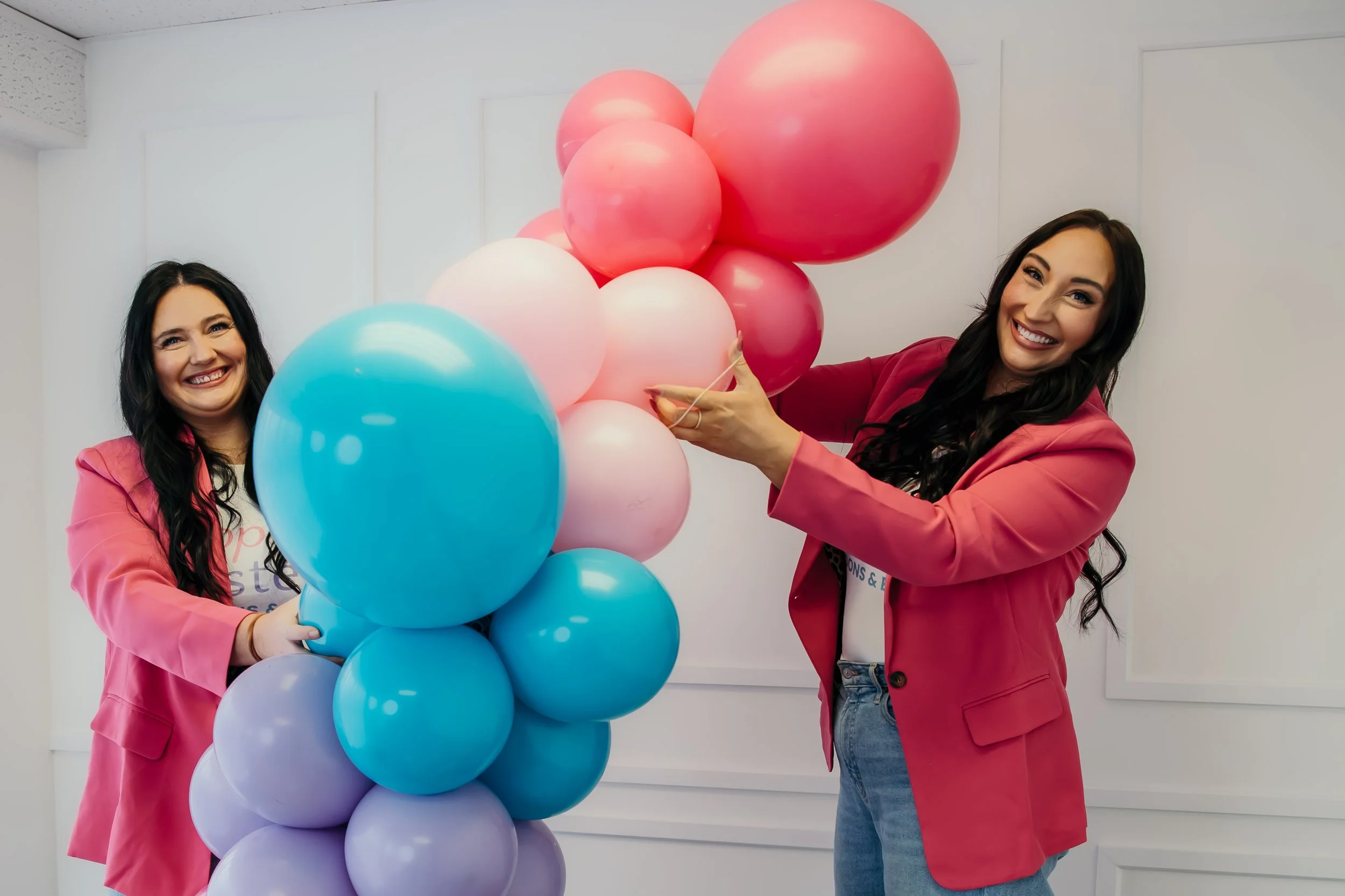 Two women in pink blazers holding a colorful balloon arch, smiling at the camera.