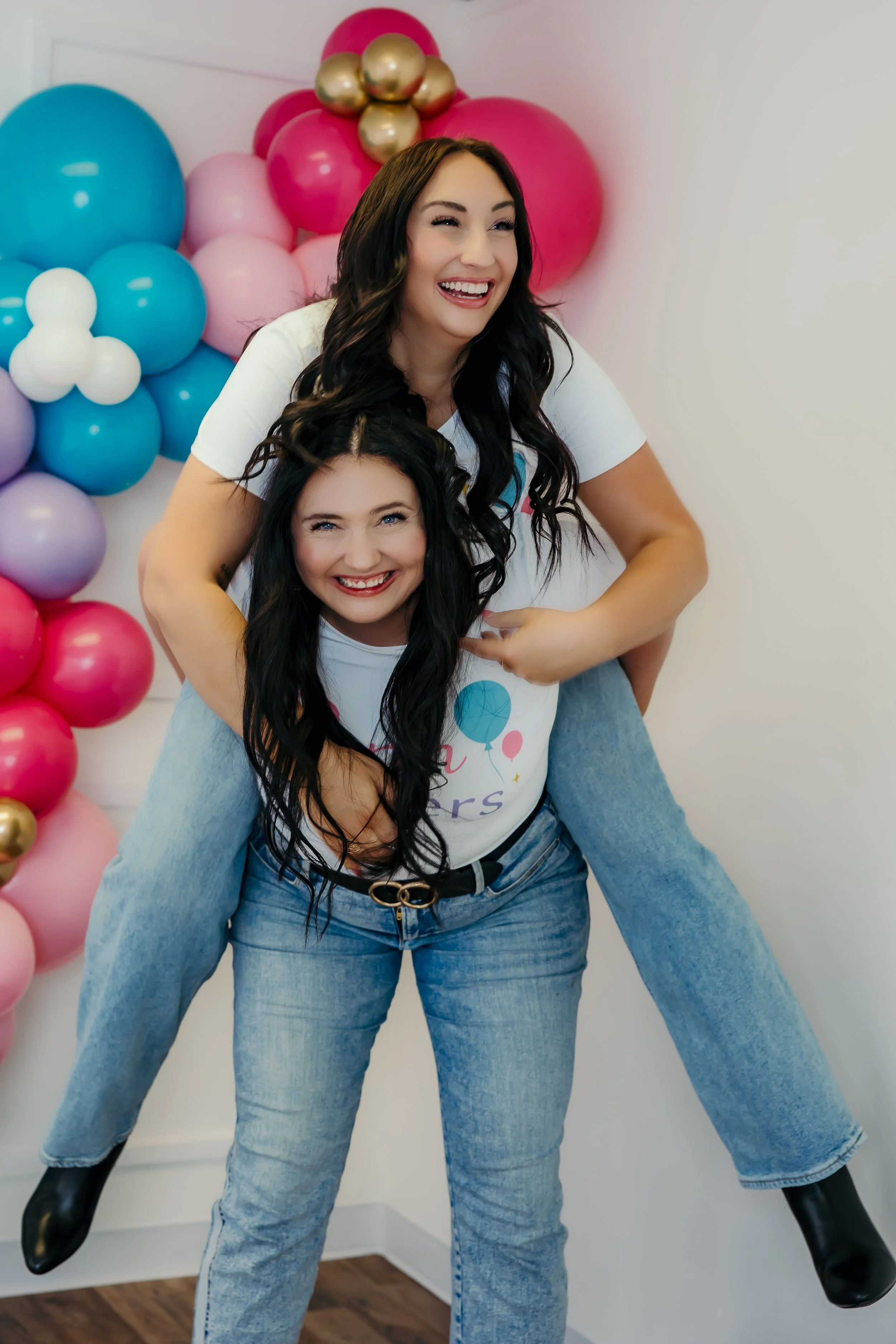 Two women smiling and having fun, with one giving a piggyback ride to the other, in front of colorful balloon decorations