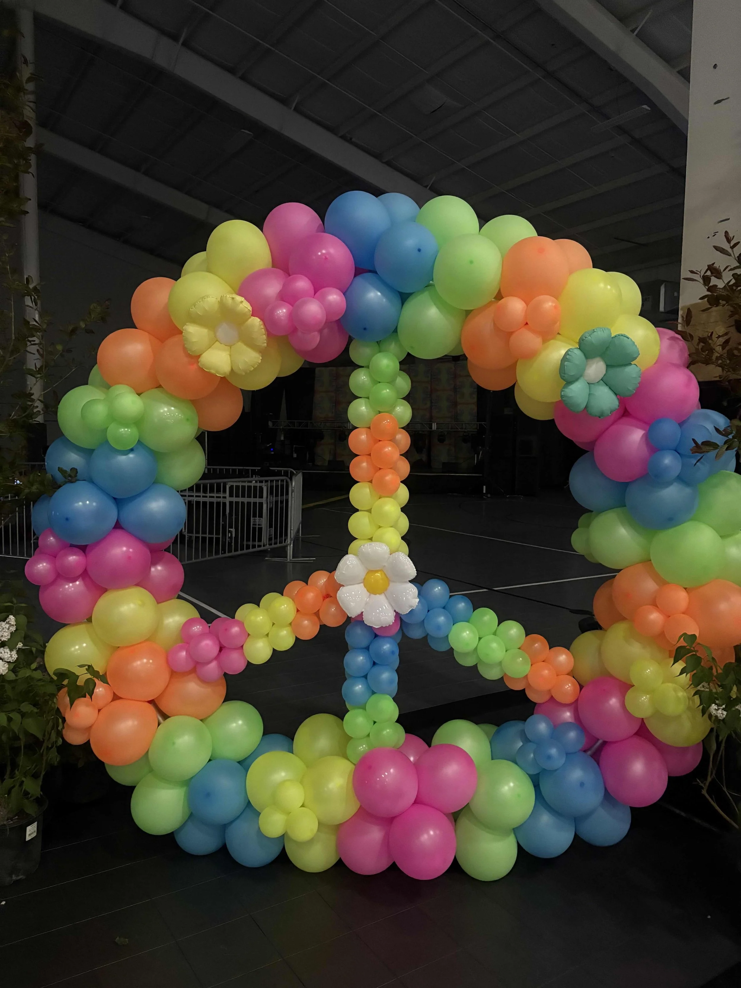Colorful balloon arrangement in the shape of a peace sign, decorated with balloon flowers, set indoors on a black floor.