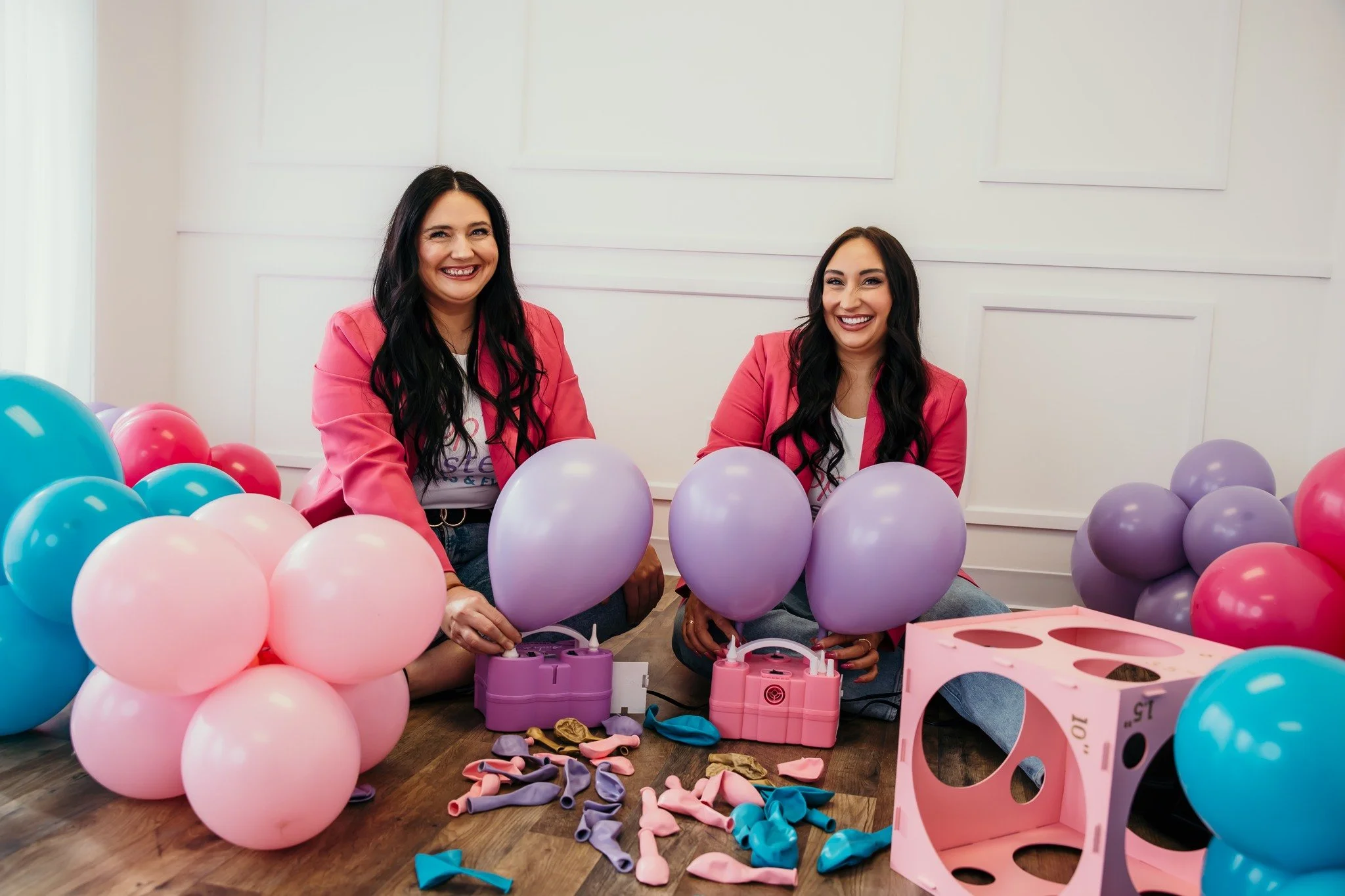 Two women smiling while preparing balloons for a celebration, sitting on the floor surrounded by pink, purple, and blue balloons and party supplies.