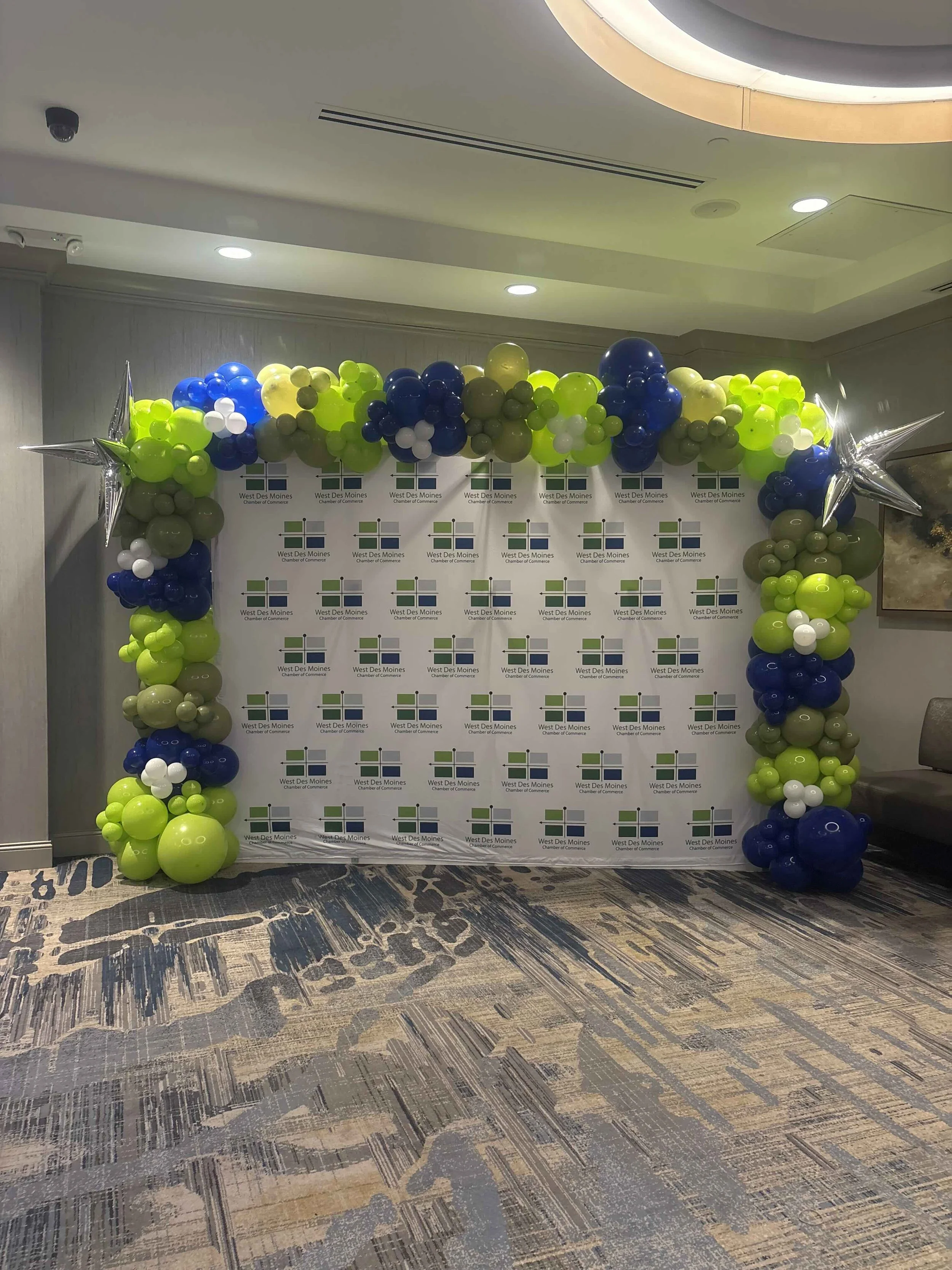 Balloon arch with green, blue, white, and gray balloons framing a backdrop with West Des Moines Chamber of Commerce logo.