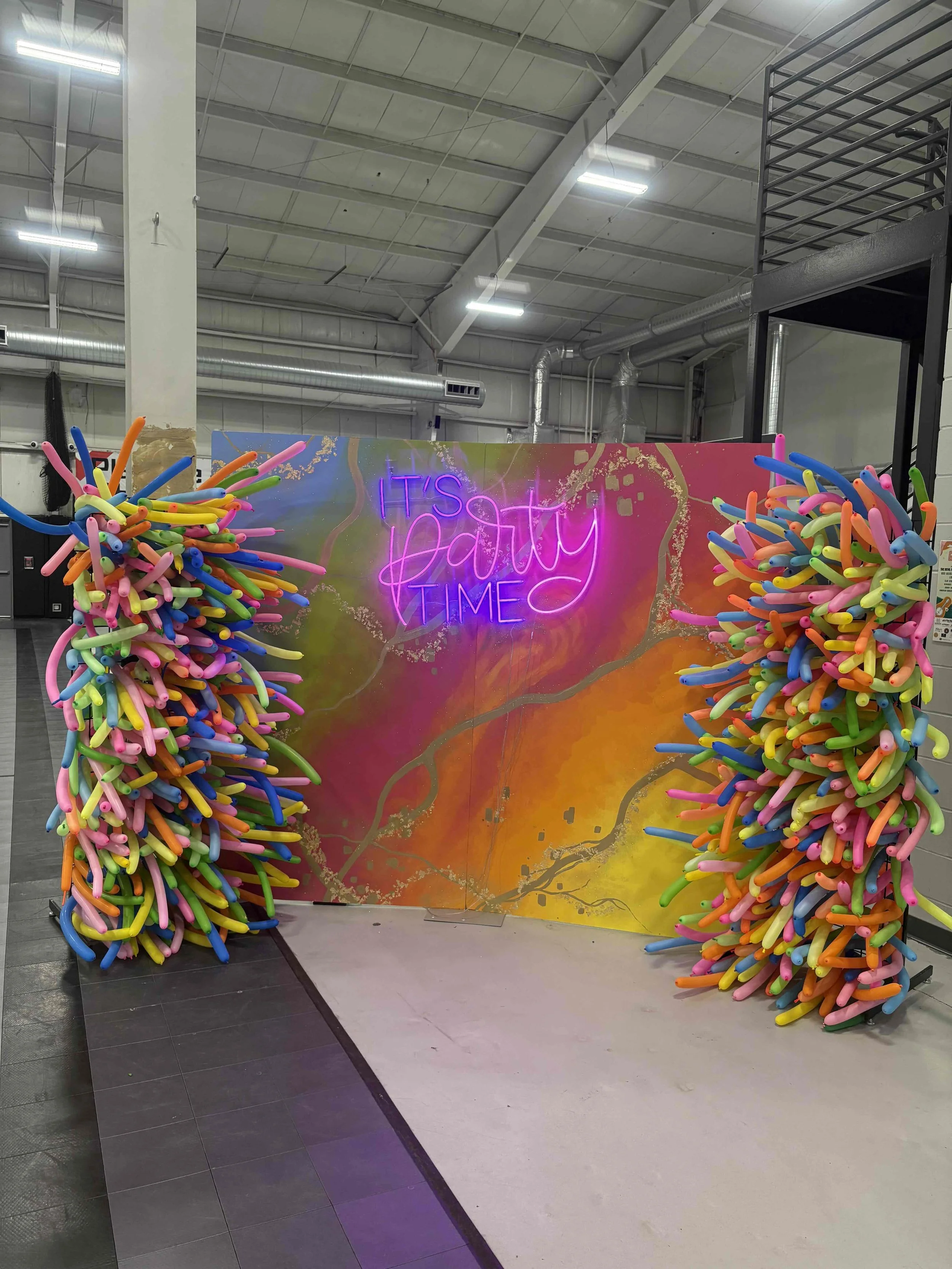 Colorful balloon arrangements on either side of a vibrant backdrop with neon sign that reads 'It's Party Time' at an indoor event space.