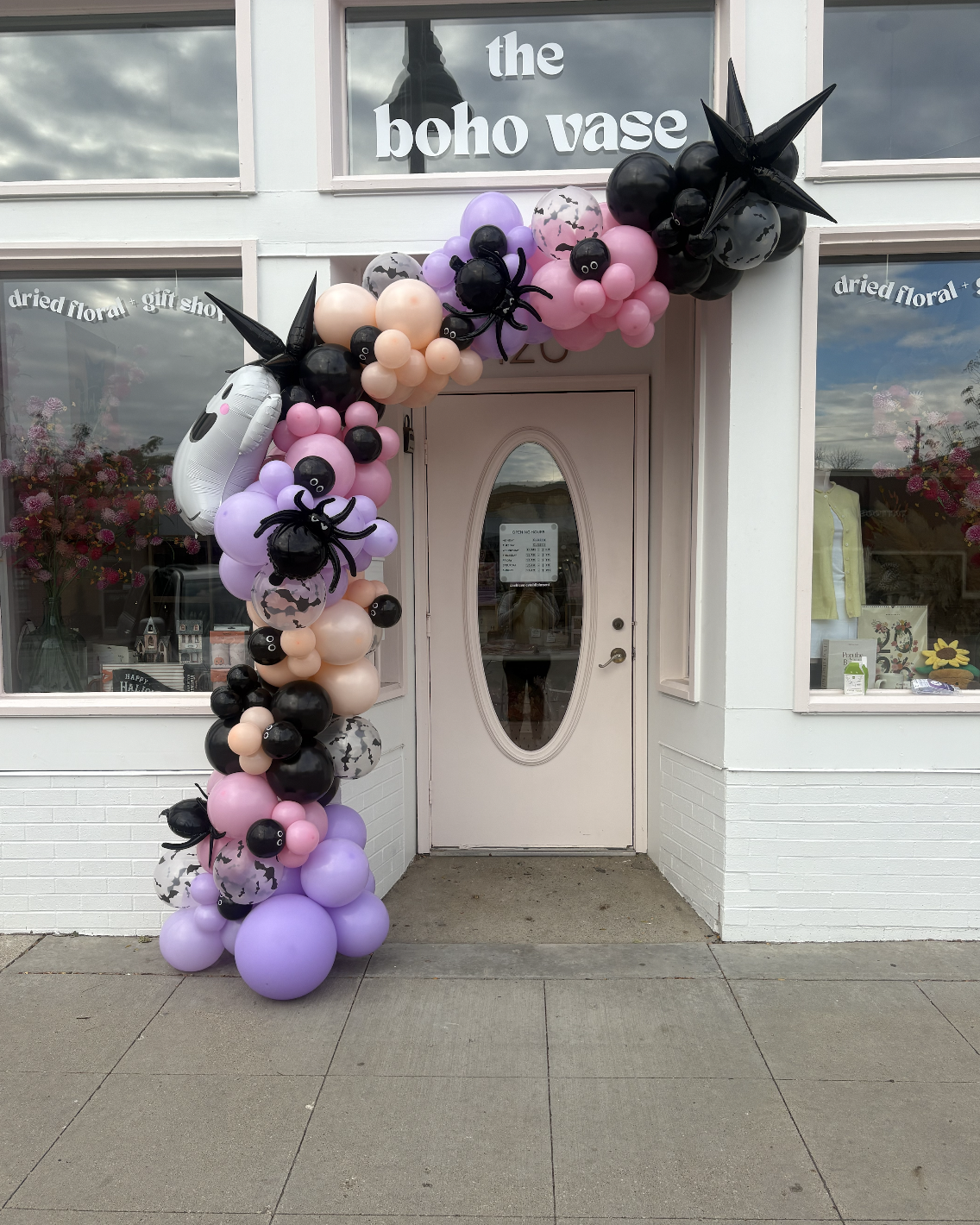 Decorative balloon arch in front of a boutique shop, featuring black, pink, purple, and marbled balloons, with some shaped like spiders and a ghost.