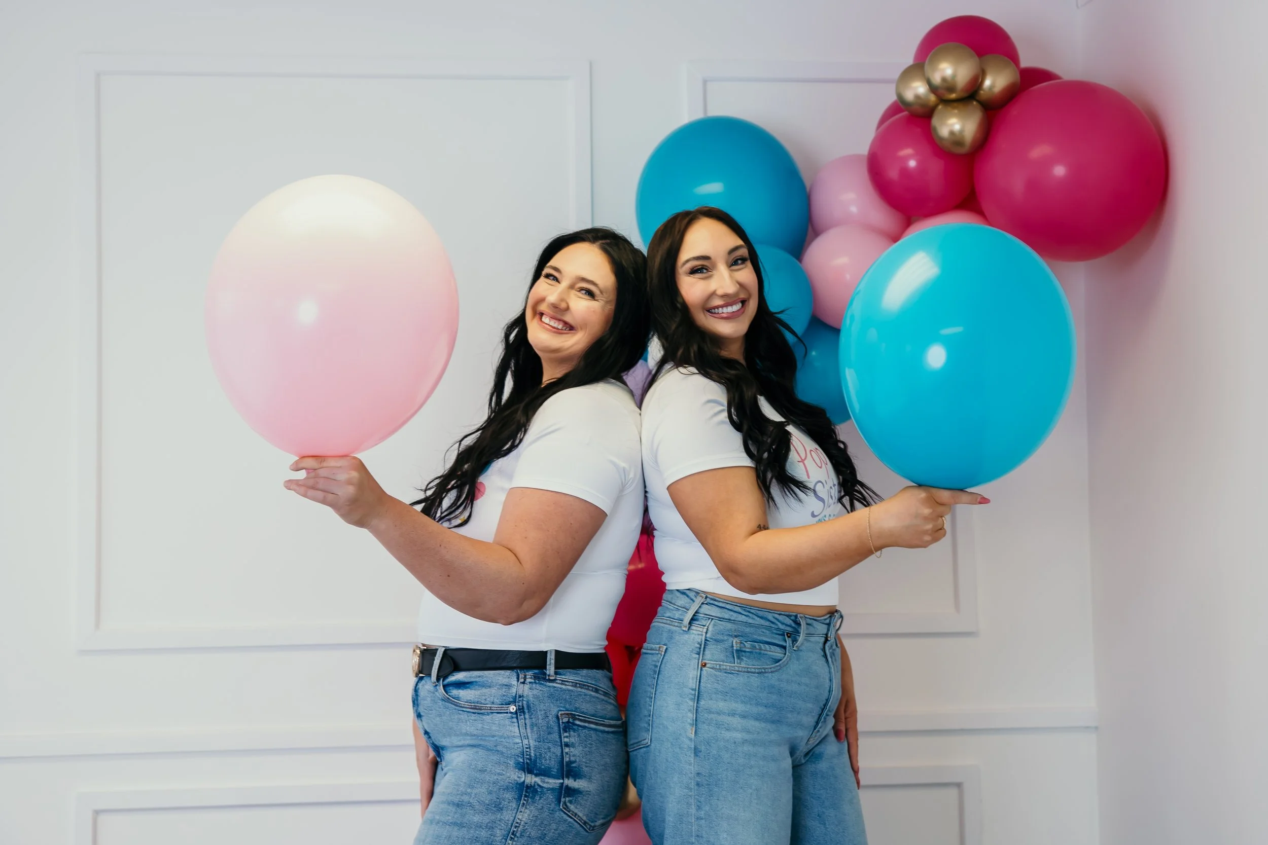 Two women standing back-to-back, holding balloons, at a celebration with pink, blue, and gold balloons in the background, smiling.