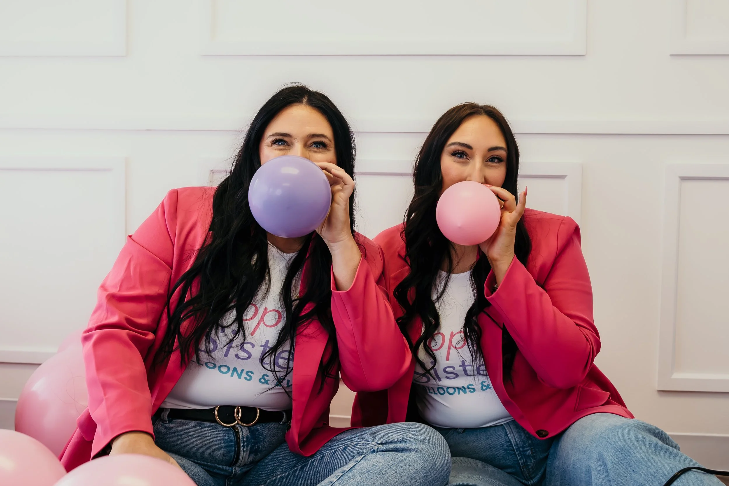 Two women wearing pink blazers and white t-shirts holding balloons in front of their faces, sitting on the floor surrounded by pink and purple balloons.