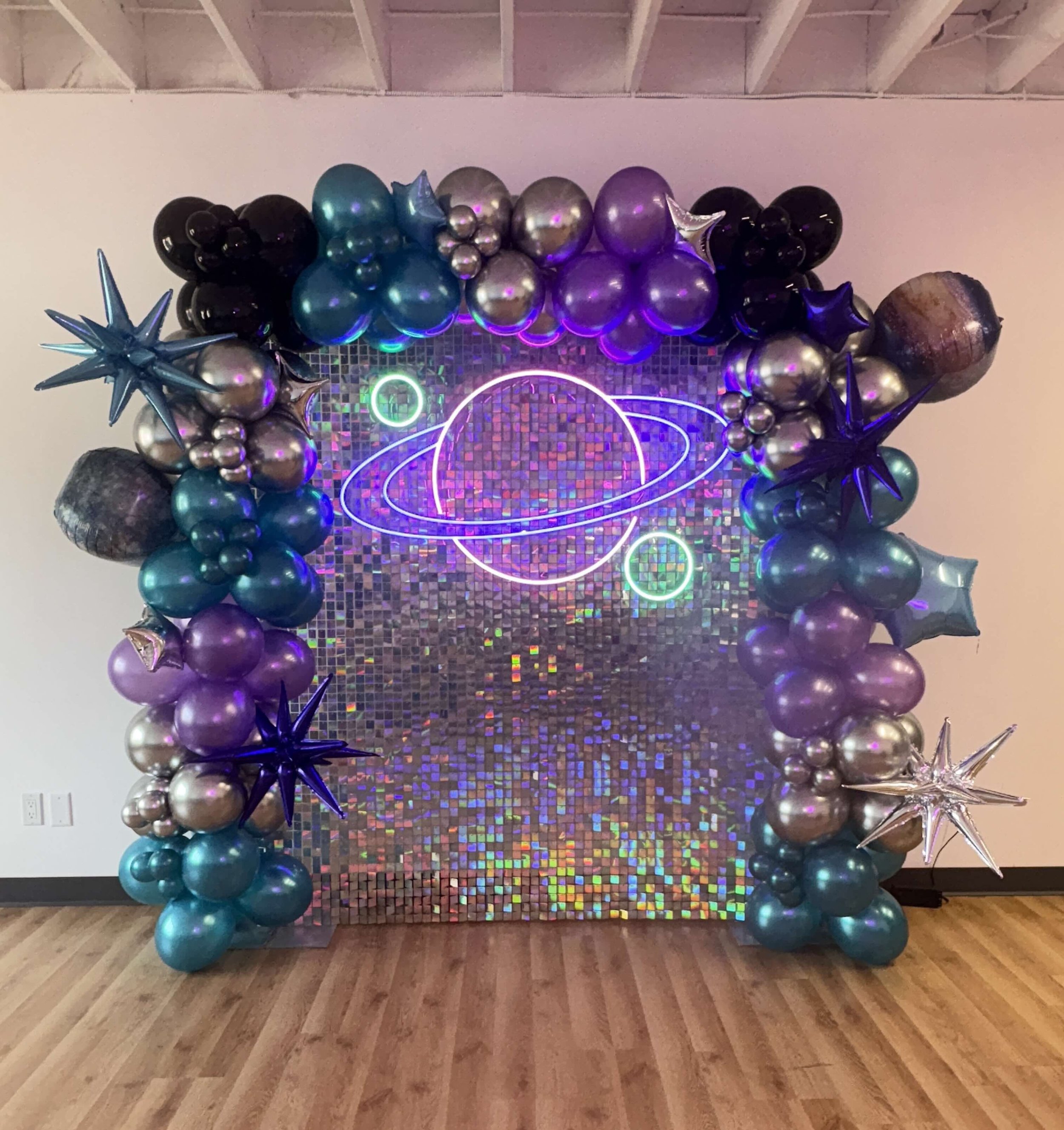 Decorative space-themed balloon arch with metallic, star-shaped, and planet-shaped balloons surrounding a holographic backdrop and neon planet and orbit design.